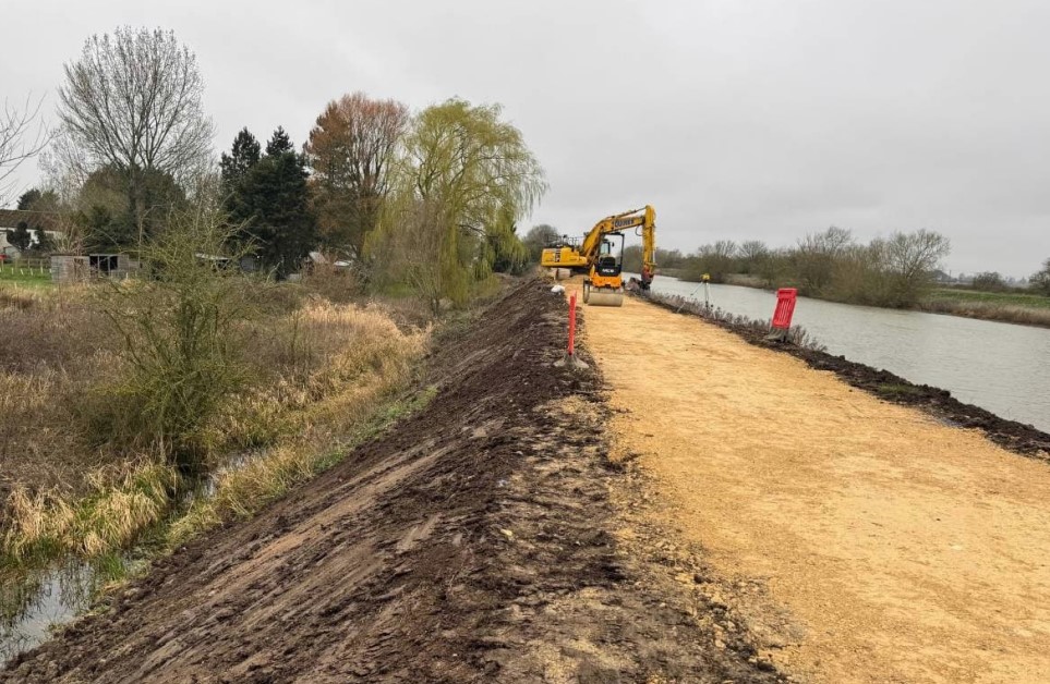 Repairs to the crest of a river embankment. A digger and a roller are on the top of the embankment in the distance carrying out work. 
