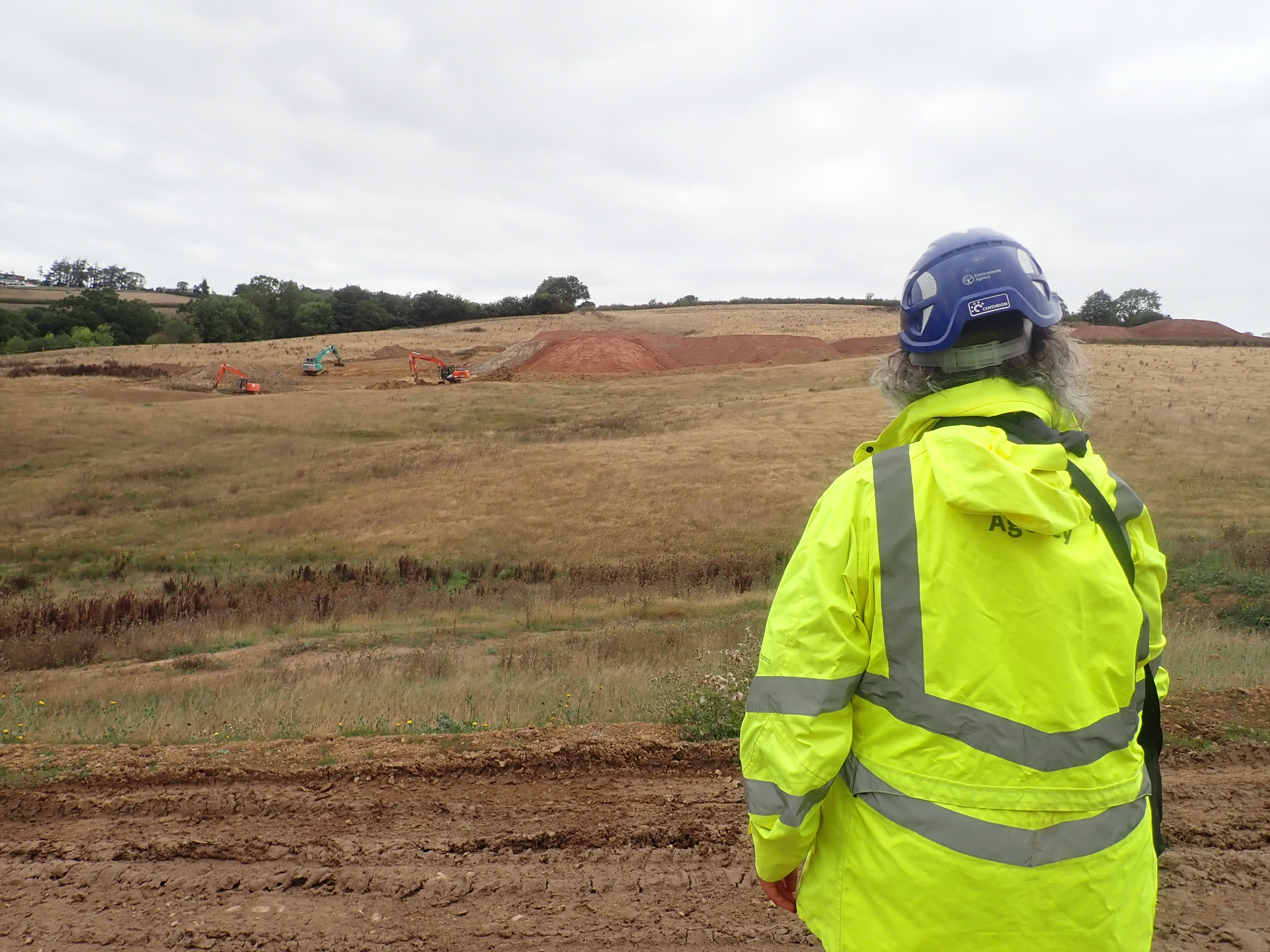 An Environment Agency officer in a hi-viz jacket stands at a distance observing diggers preparing a grassy field to become a landfill site.