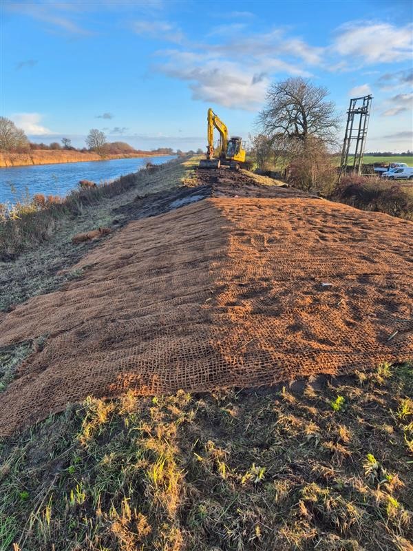A raised river embankment with a geotextile mesh covering bare earth where a low spot has recently been raised. There is a small digger in the background. 