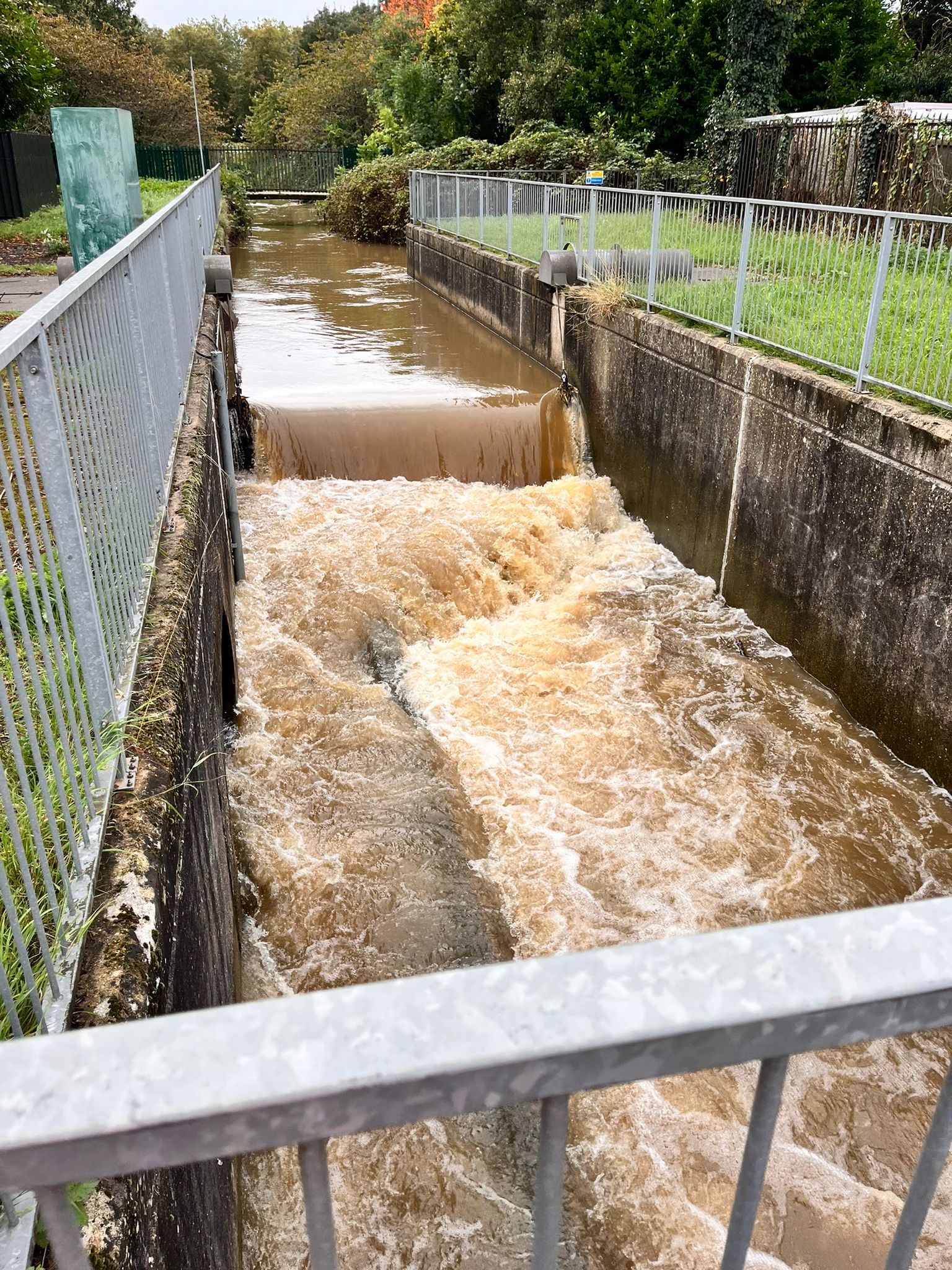 A photo looking down into and along a concrete channel several metres wide and a few metres deep below the surrounding ground level. Galvanised fencing lines the channel edge. The channel is two thirds full of brown flowing water, which is flowing over a gate, which isn't visible due to the water, forming a water fall about 50 centimetres high and creating turbulent, white aerated flows downstream.