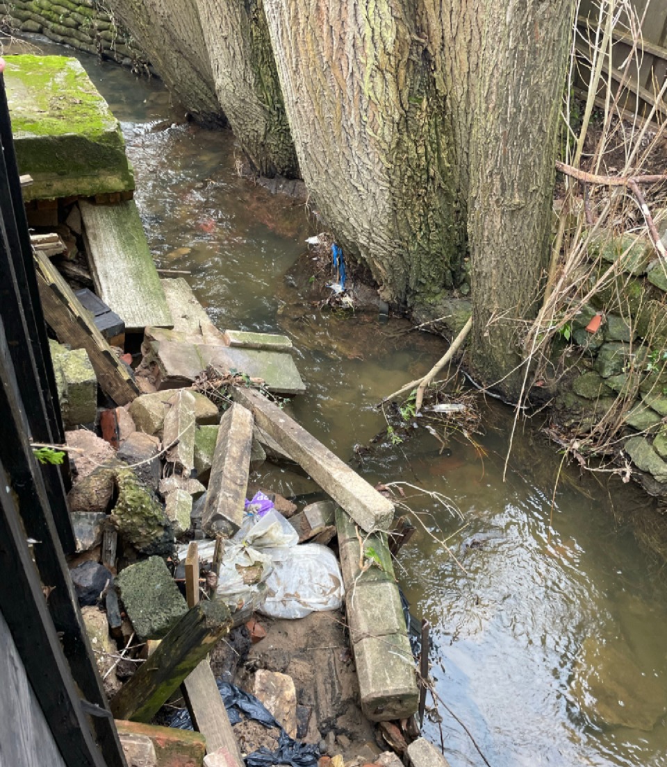 A brook about 2 meters wide, with water about 20centimetres deep. Half of the channel width is taken by an assorted pile of debris/waste, mostly concrete blocks and rubble.