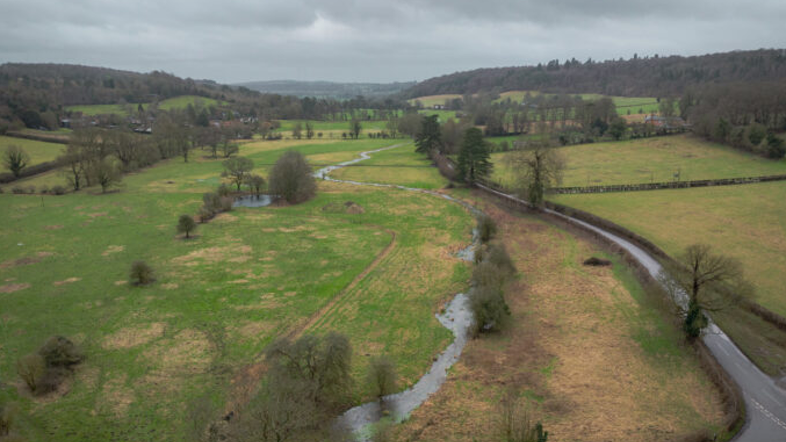 Aerial view of a rural landscape with green fields, a winding stream, scattered trees, and a road. Overcast sky.