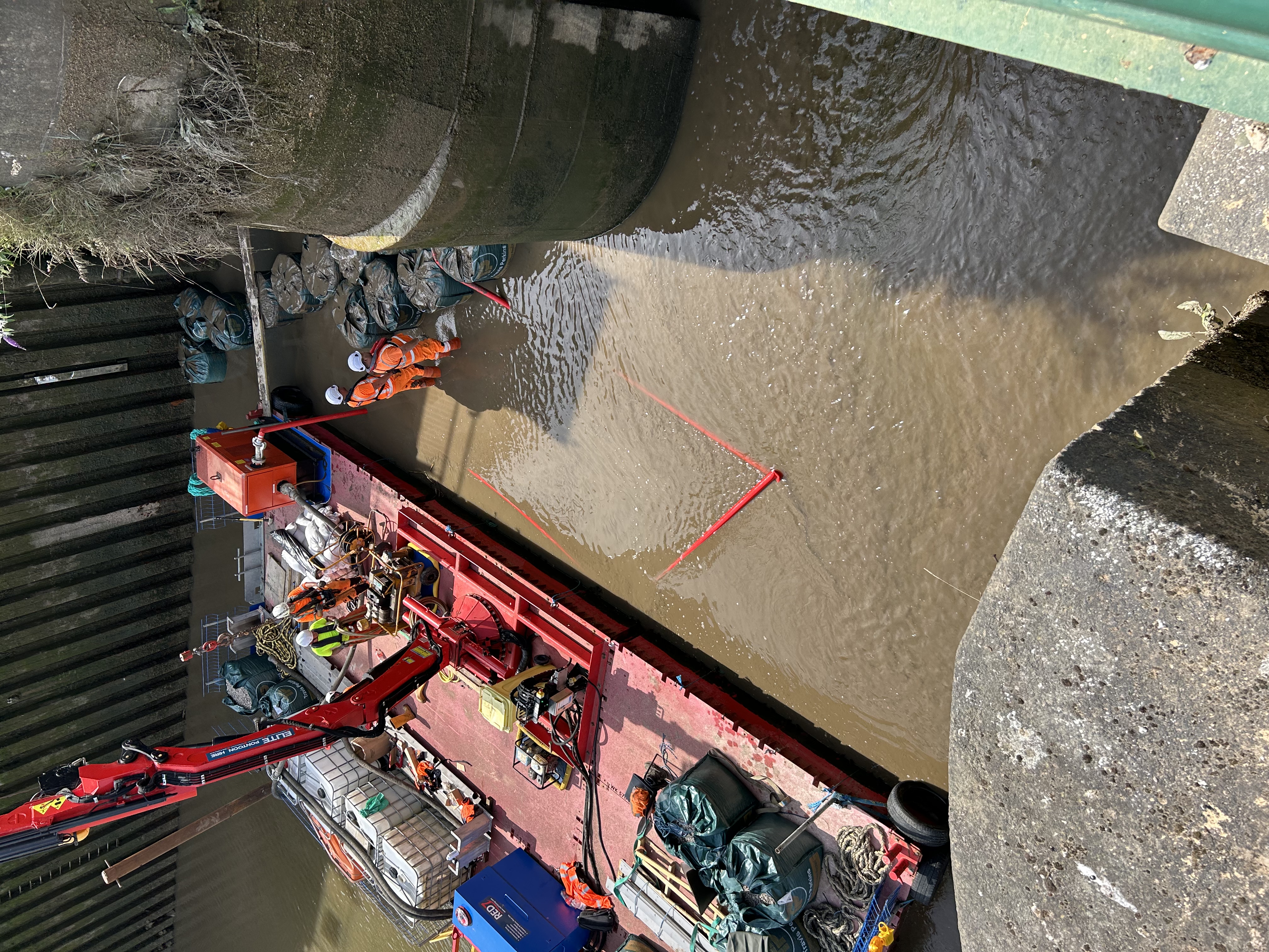 Workers in orange hi vis clothing stand in shallow water in the channel at Grand Sluice. They are beside a red barge with a crane that they are using to carry out works on the structure. 