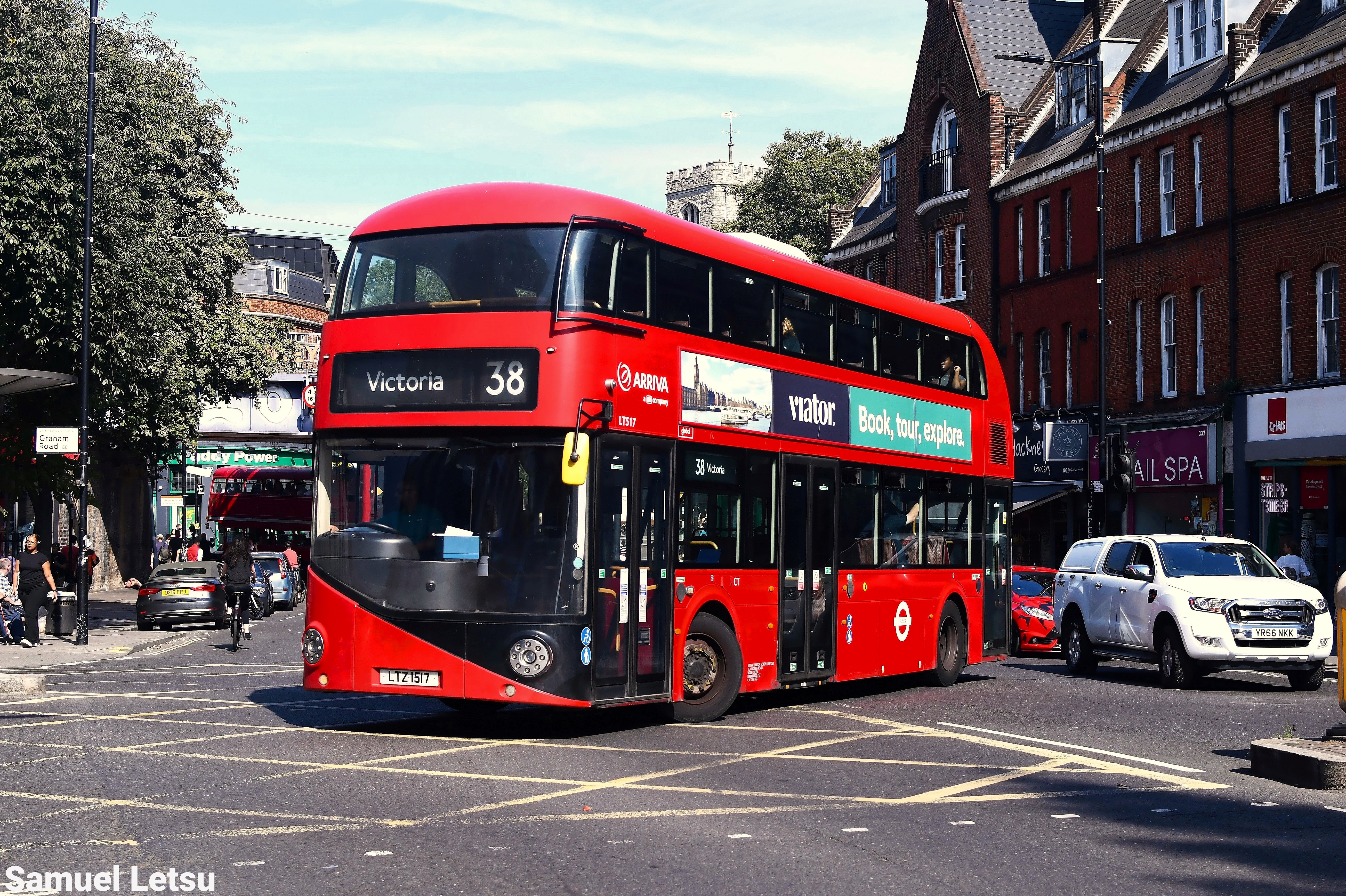 Red double-decker London bus on route 38 to Victoria, turning at an urban street junction with cars and brick buildings in the background