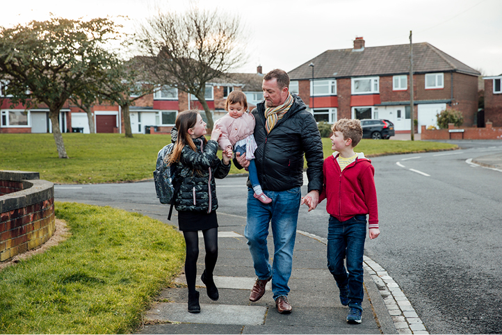 A family of four (including three children and a man) walking along a street holding hands. The man is holding a small child.