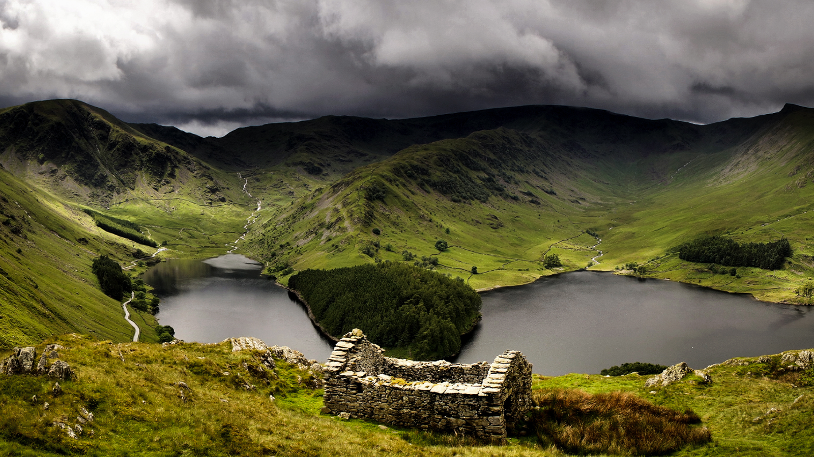 A panoramic view of lush green hills surrounding two lakes under a dramatic, cloud-filled sky, with a stone ruin in the foreground.