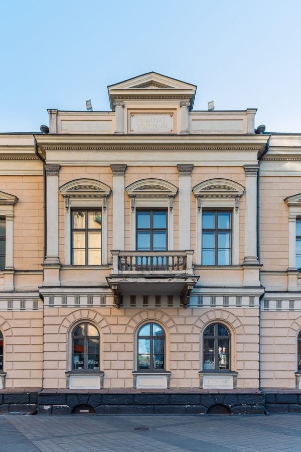 A photo of an old stone building with two floors and a balcony in the middle. The windows have arches. The building is light yellow with creamy white on pillars and details. There's an old stone plate with the year 1858 engraved in gold Roman numbers at the top of the building above the balcony.