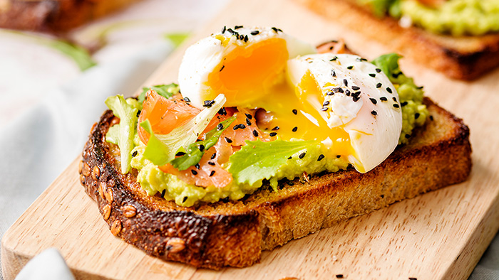 Geräucherter Lachstoast mit Avocadocreme und pochiertem Ei, aus dem cremiges Eigelb tropft. /	mauritius images / The Picture Pantry / Massimo Aggius Vella