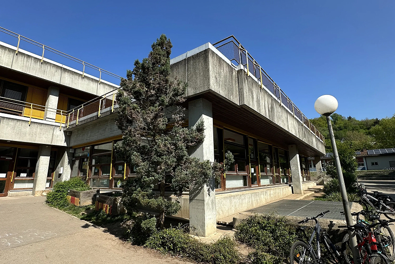 Betonbau mit großen Fenstern, umgeben von Pflanzen und Fahrrädern.