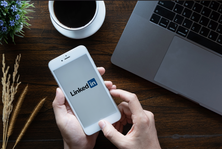 Arbeitsplatzansicht: Links steht eine Blumendeko, oben eine Kaffeetasse. Rechts im Bildausschnitt ein Laptop, davor zwei Hände, die ein Smartphone halten. Auf dem Display ist die LinkedIn-App geöffnet.