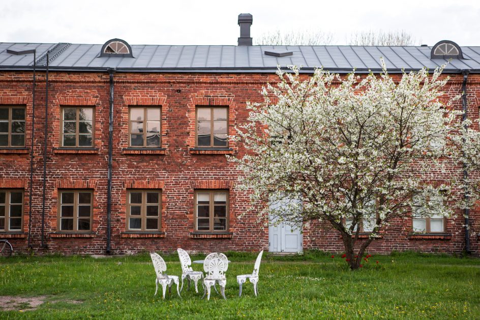 Kuvassa tiilitalo, kukkiva omenapuu ja valkoiset puutahakalusteet.