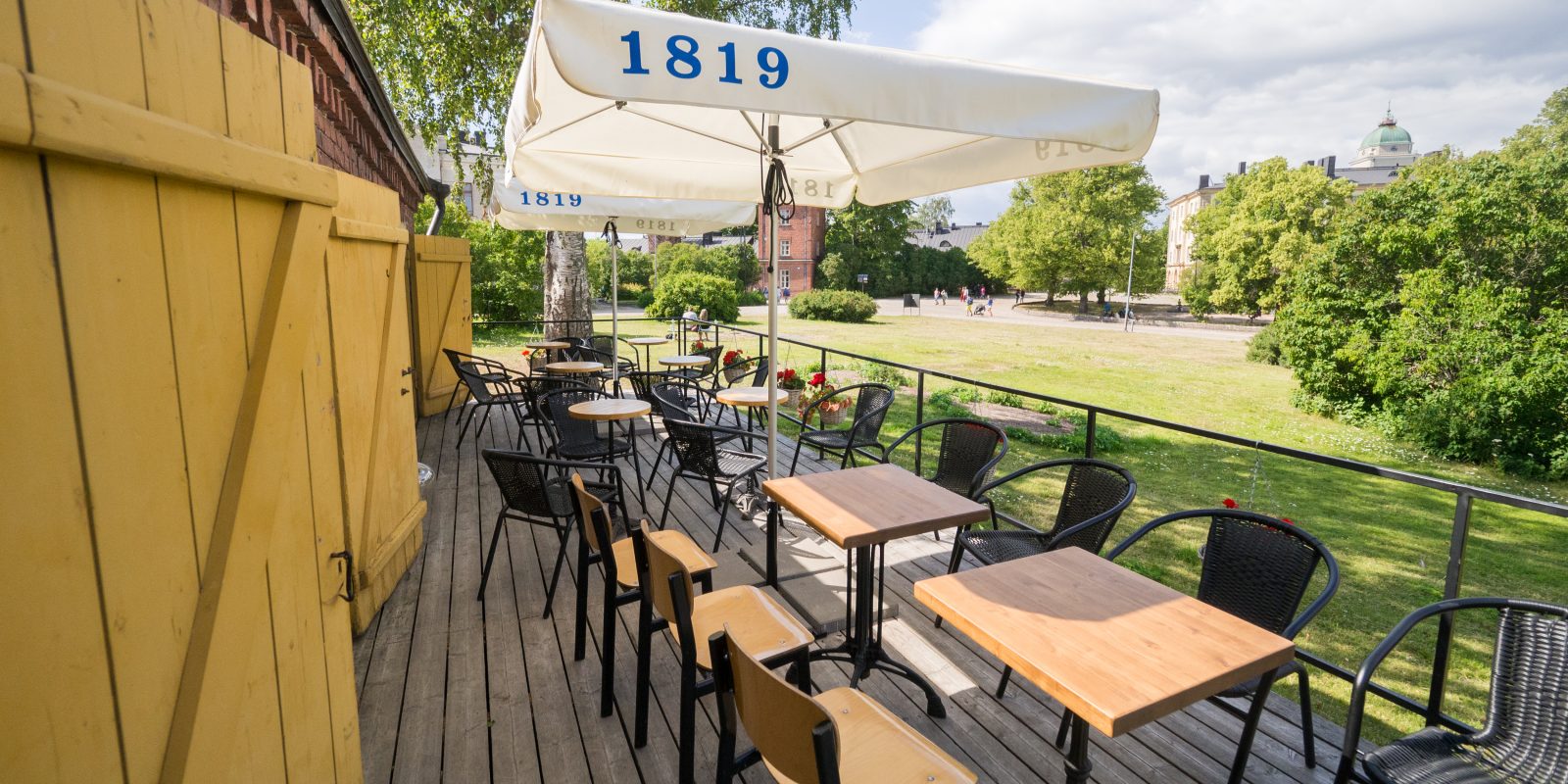 Kuvassa Linna baarin terassi, jossa tuoleja, pöytiä ja punaisia kukkia. Taustalla puistoa ja taivasta
