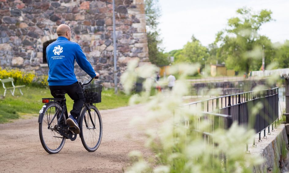 Kuvassa Suomenlinnan kesävalvoja pyöräilee. Hänellä on sininen paita jossa valkoisella teksti suomenlinna ja logo. Taustalla muuria ja etualalla valkoisia kukkia.