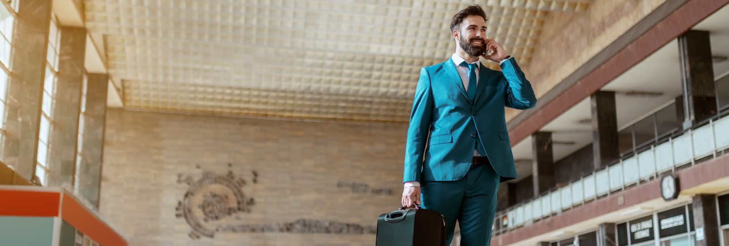 Young attractive bearded smiling businessman in blue suit carrying luggage and using smart phone while standing at train station. business trip concept