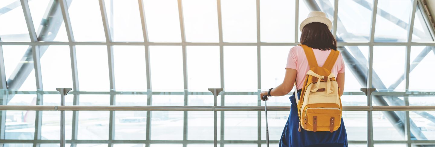 Asian young woman with suitcase and yellow backpack waiting for the flight at window of the airport