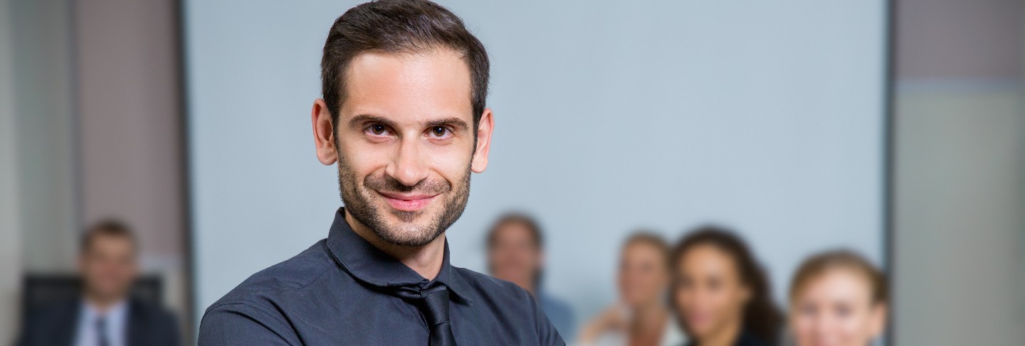 Man smiling with suit sitting at a table with colleagues behind
