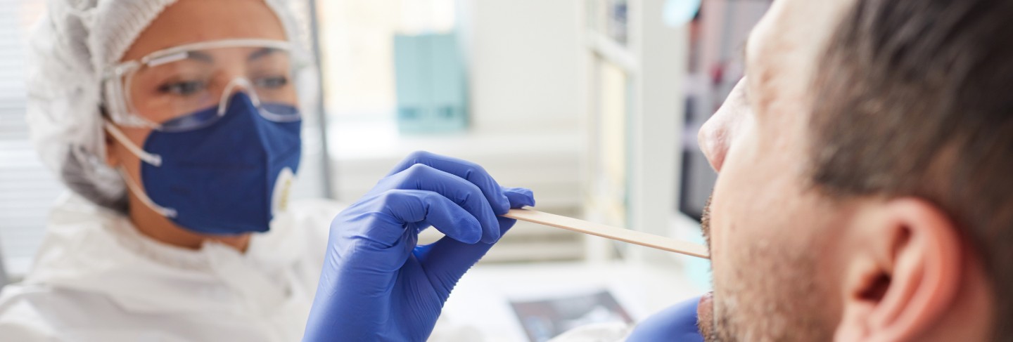 Mature man opening the mouth while doctor in protective clothing examining his throat during medical exam at hospital
