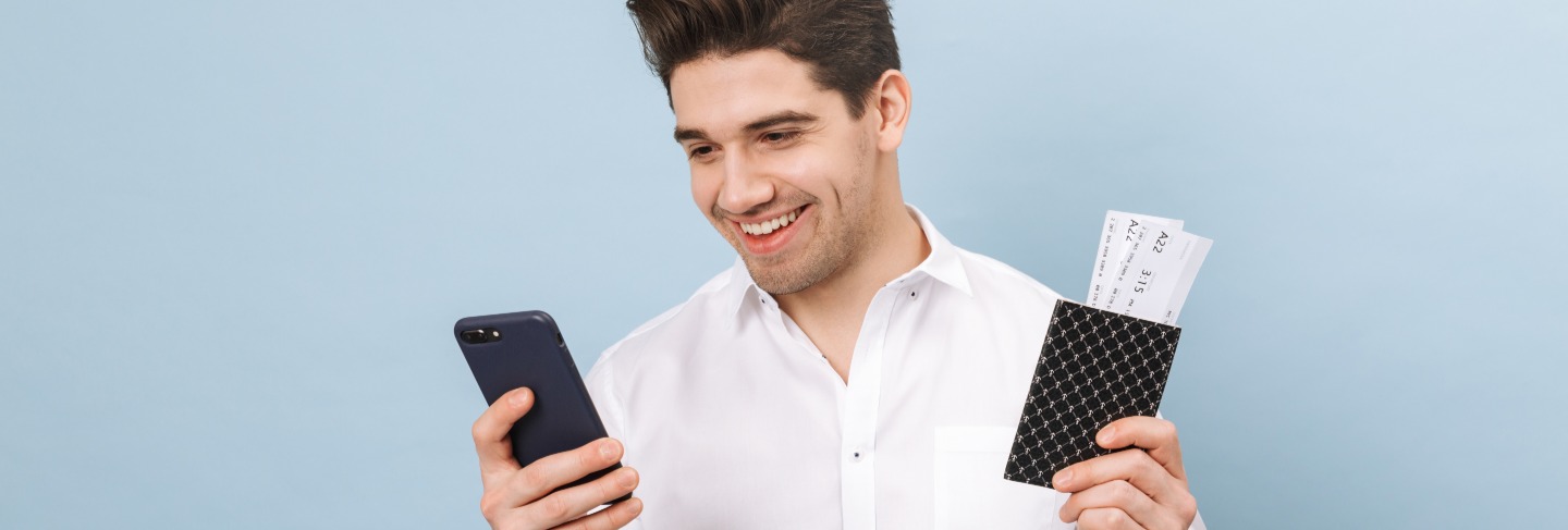 Portrait of a cheerful handsome young man standing isolated on blue, holding passport with flight tickets, using mobile phone