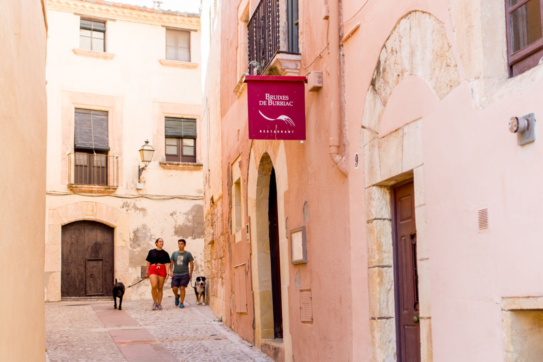 Una pareja y sus perros pasean junto al restaurante 'Bruixes de Burriac', en Carrer del Cup. Una pareja y sus perros pasean junto al restaurante 'Bruixes de Burriac', en Carrer del Cup.