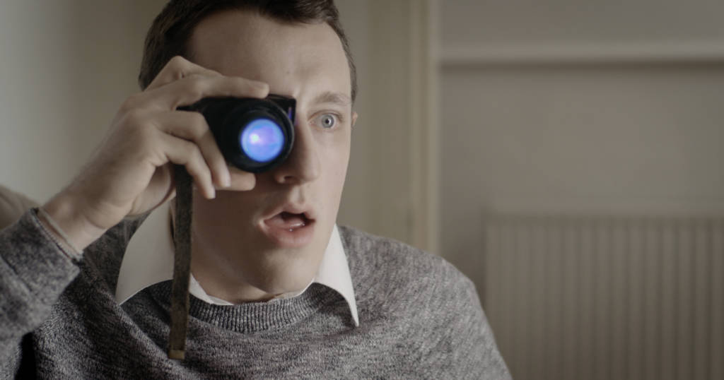 White man holds a lens up to his eye with an amazed expression