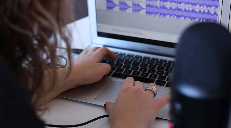 A person recording a podcast on a laptop, a microphone stands in the foreground.