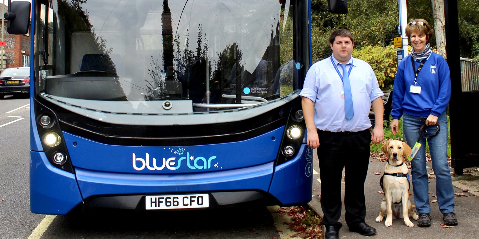 Bluestar's George Miller with trainer Jane Tabor and Guide Dog Poppy stand next to the front of a Bluestar Bus