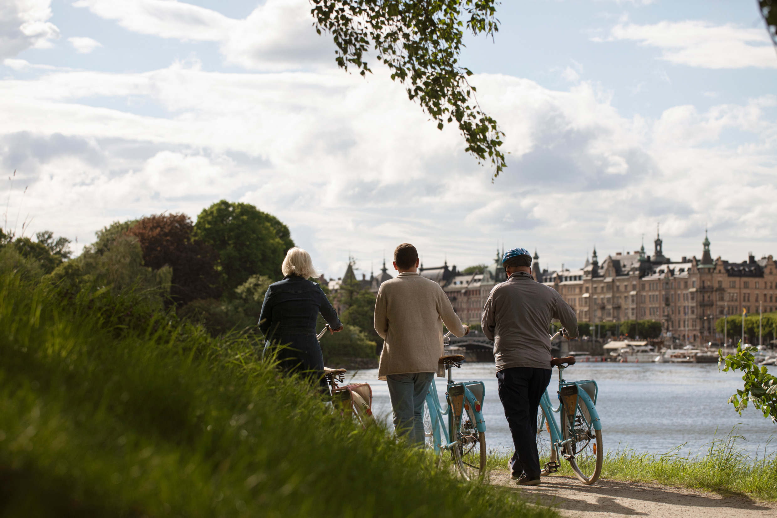 Tre personer står och håller i sina cyklar medan de tittar ut över vattnet med slottsliknande byggnader i bakgrunden och grönska omkring dem.