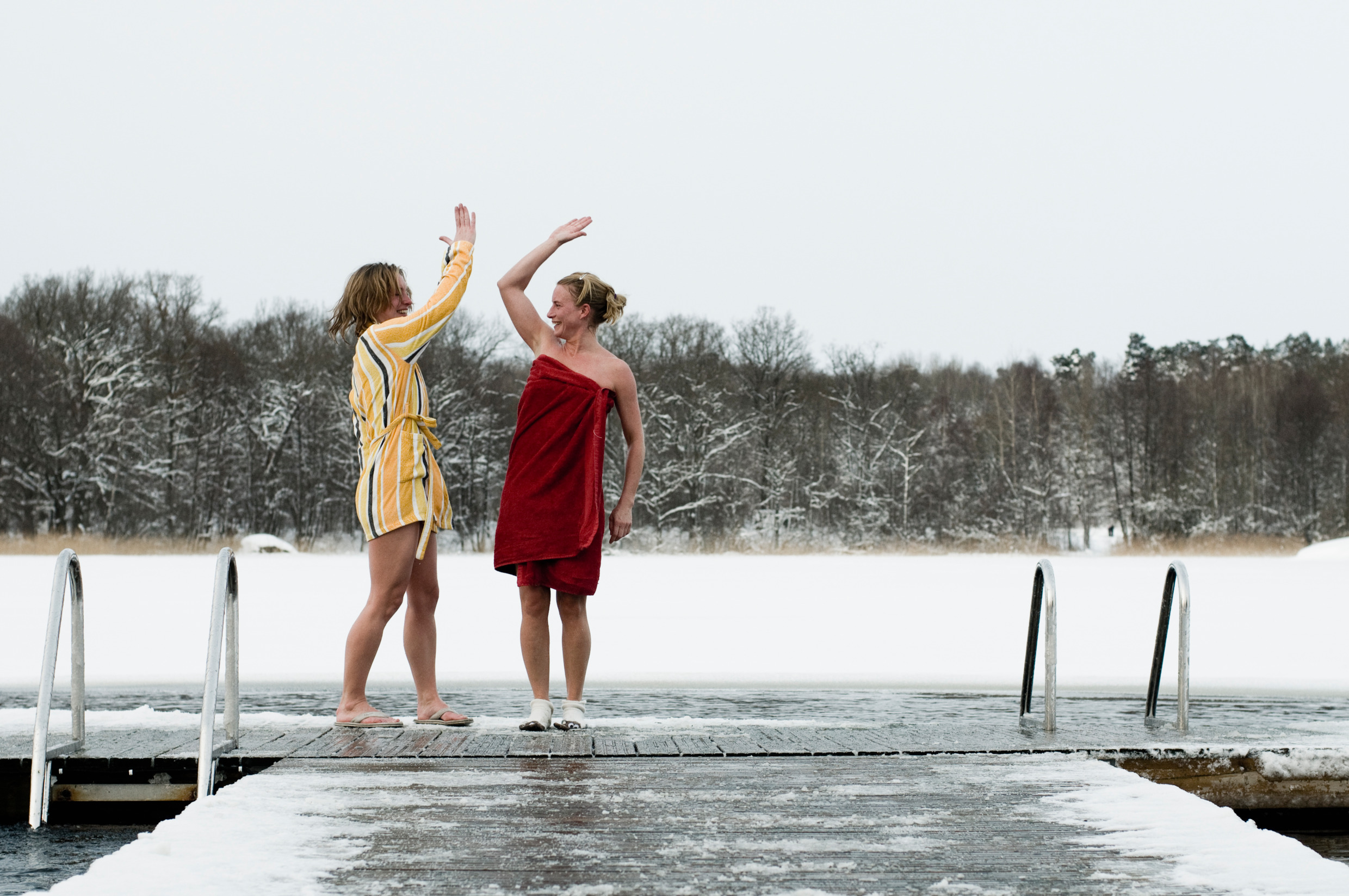 Two women are high-fiving on a dock by the water. It is winter and one is wearing a red towel and the other one is wearing a bathrobe.
