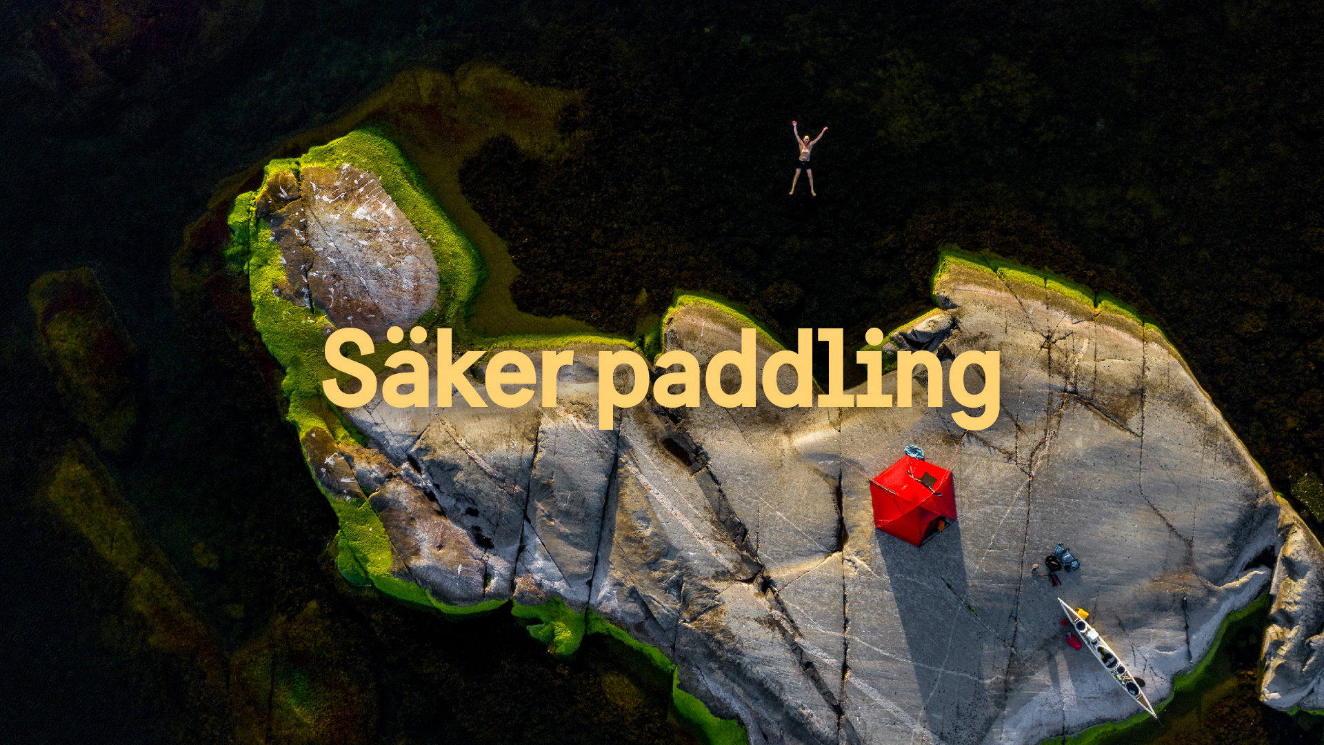 En bild tagen ovanifrån på en liten kobbe i skärgåden. På kobben syns ett tält och en kajak. En person ligger i vattnet raklång och badar och njuter.