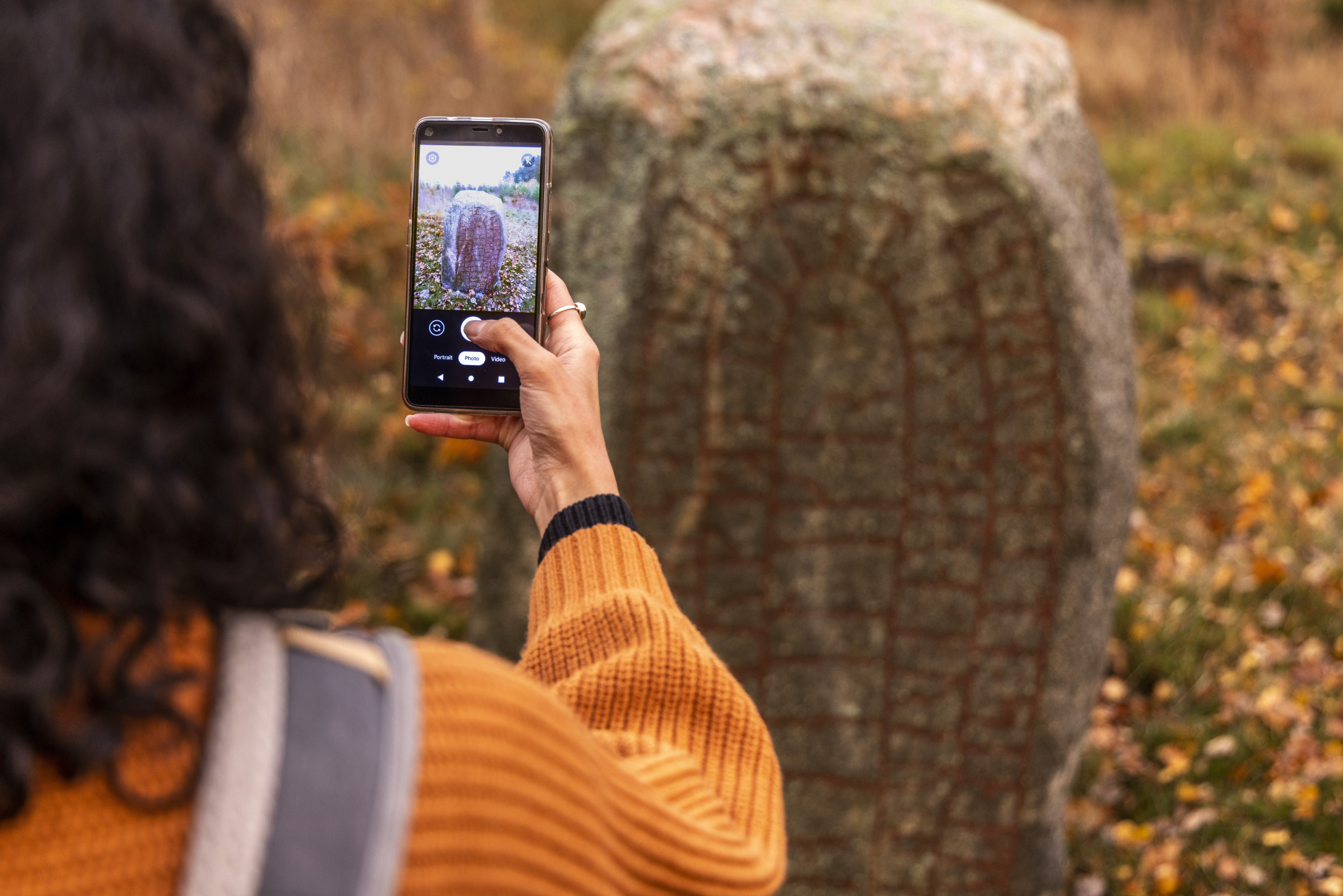 En person tar kort på en runsten i naturen med en mobilkamera.