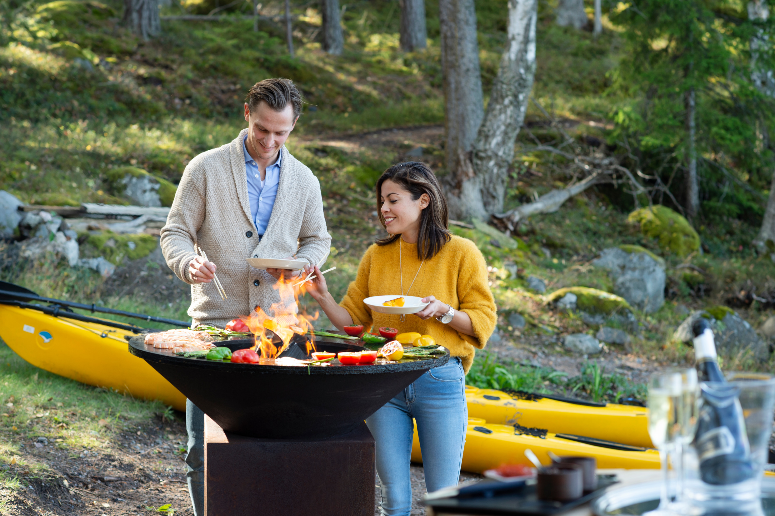 Två personer står och lagar mat på en stor wokpanna i naturen. I bakgrunden ligger ett flertal kajaker uppdragna på stranden.