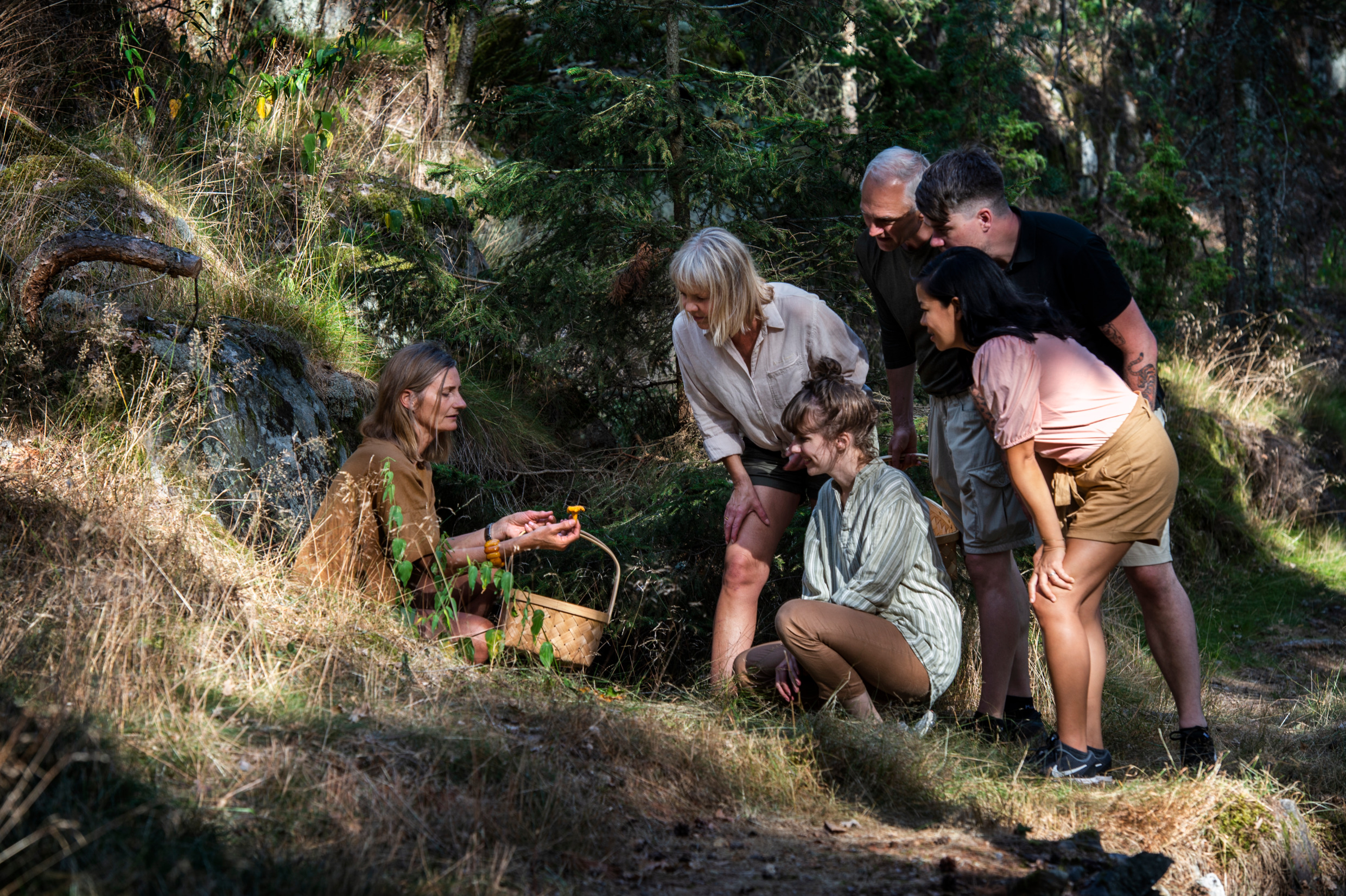 En kvinna i skogen håller i en korg och visar en kantarell för fem personer som står framåtlutade och tittar i hennes hand.