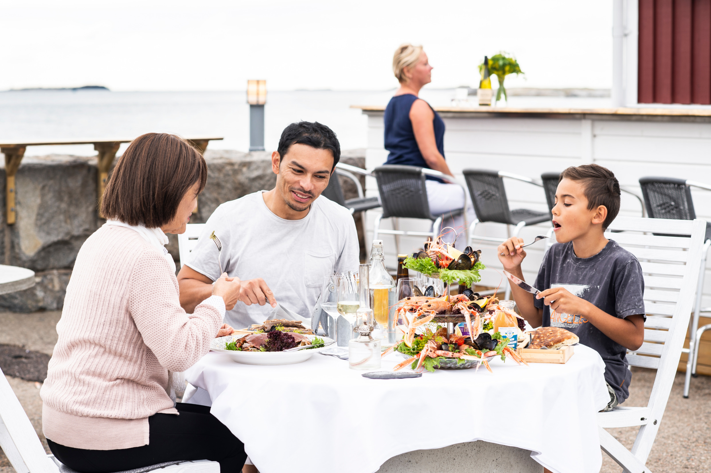 En sällskap bestående av tre personer i olika åldrar sitter och äter skaldjur till middag vid ett runt bord på en utomhusrestaurang i skärgården.