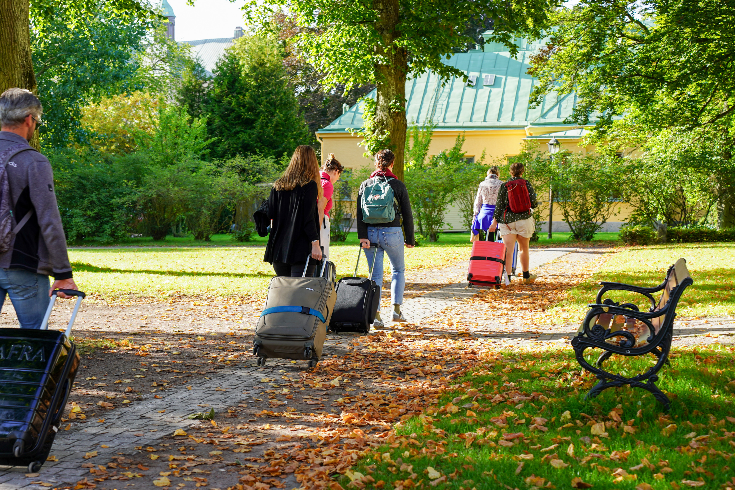 En utomhusbild föreställande 6 personer som drar varsin väska i en grusgång på väg mot en herrgårdsliknande byggnad.