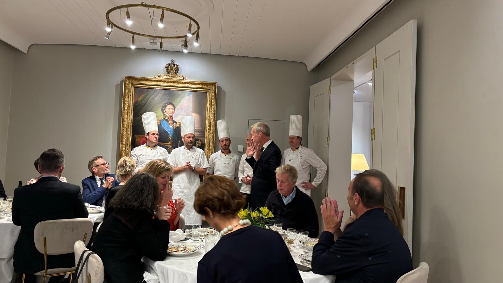 Fem kockar pratar och presenterar mat som serverats till två runda bord fyllda med personer som förväntansfullt tittar och lyssnar.