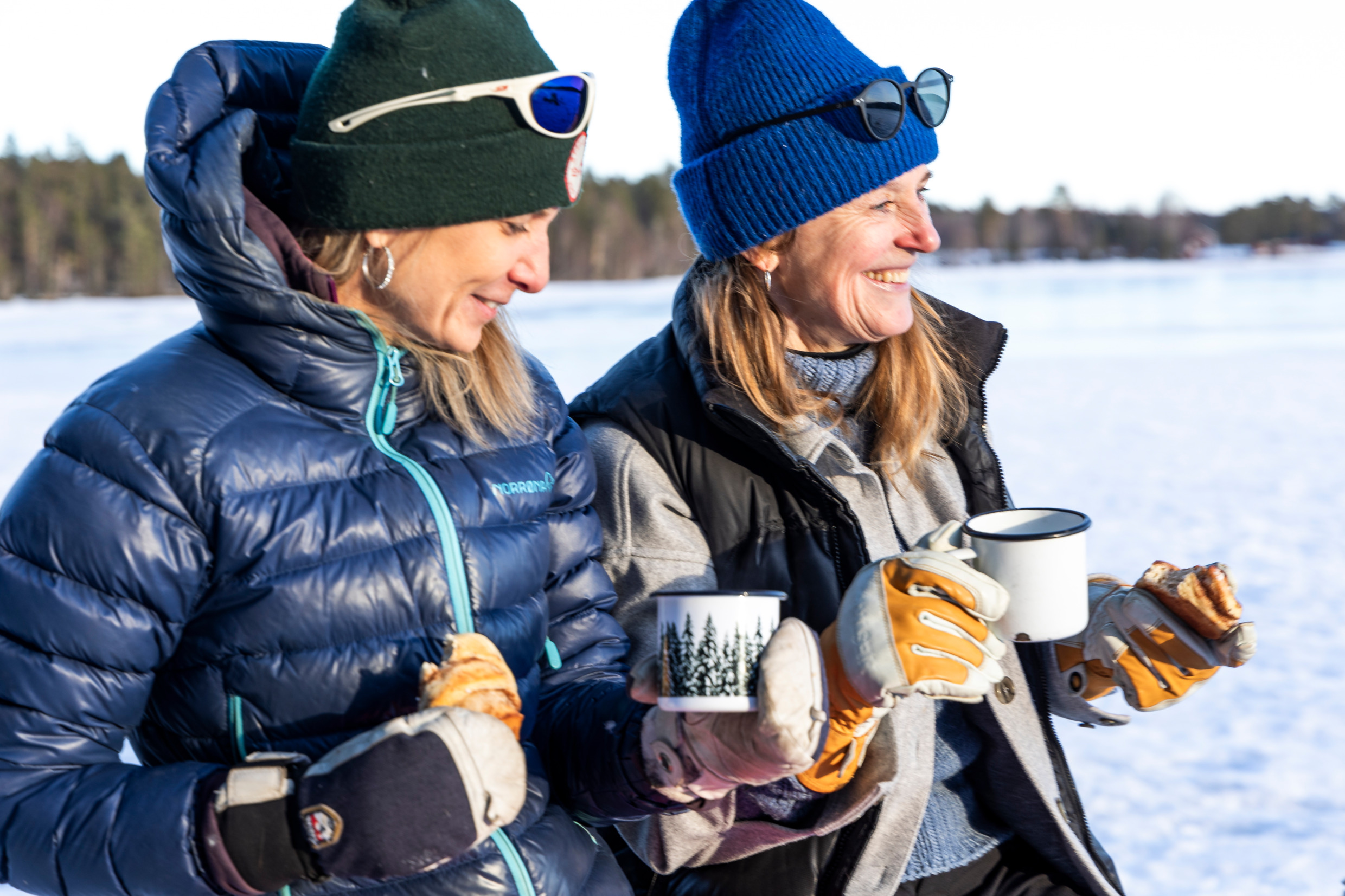 Två personer äter en varsin bulle till en kopp kaffe ståendes utomhus på en is i solen.
