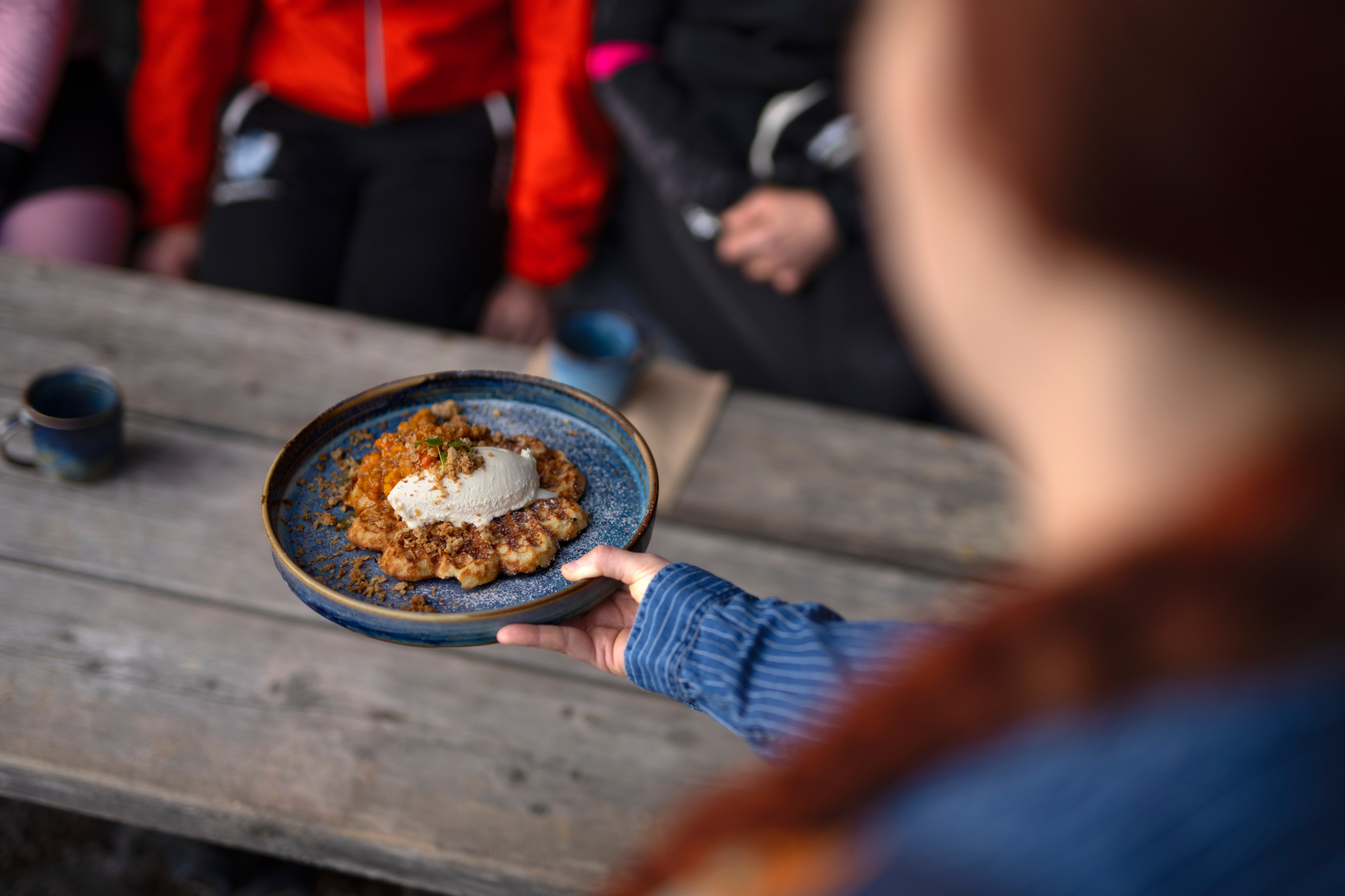 En hand håller fram en tallrik med en nygjord våffla på en fin tallrik till några personer som sitter och väntar på mat vid ett träbord utomhus.