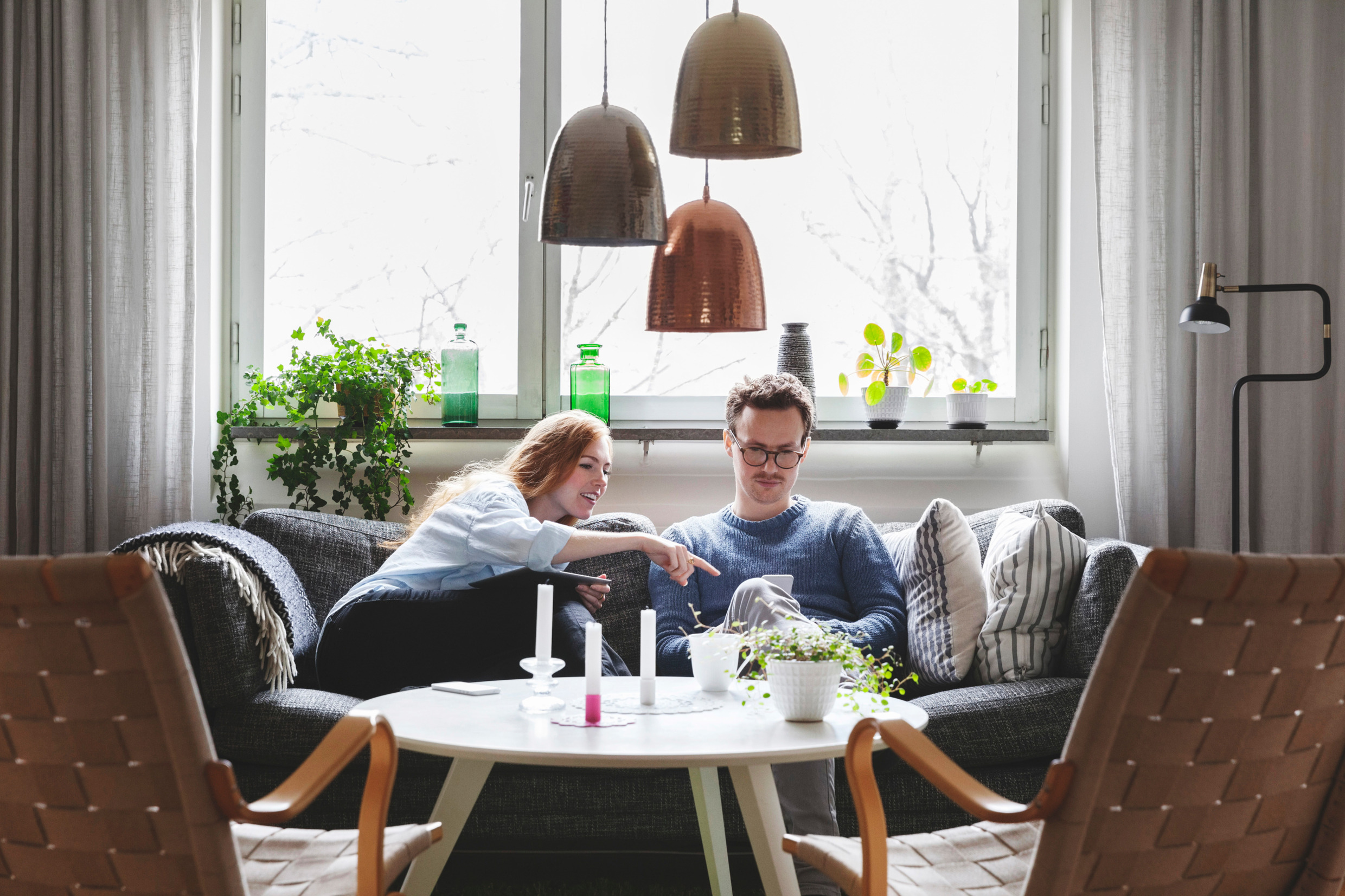 En kvinna och en man sitter i en soffa i ett vardagsrum. Kvinnan pekar på en mobiltelefon som mannen håller i handen. Kvinnan har en surfplatta i handen.