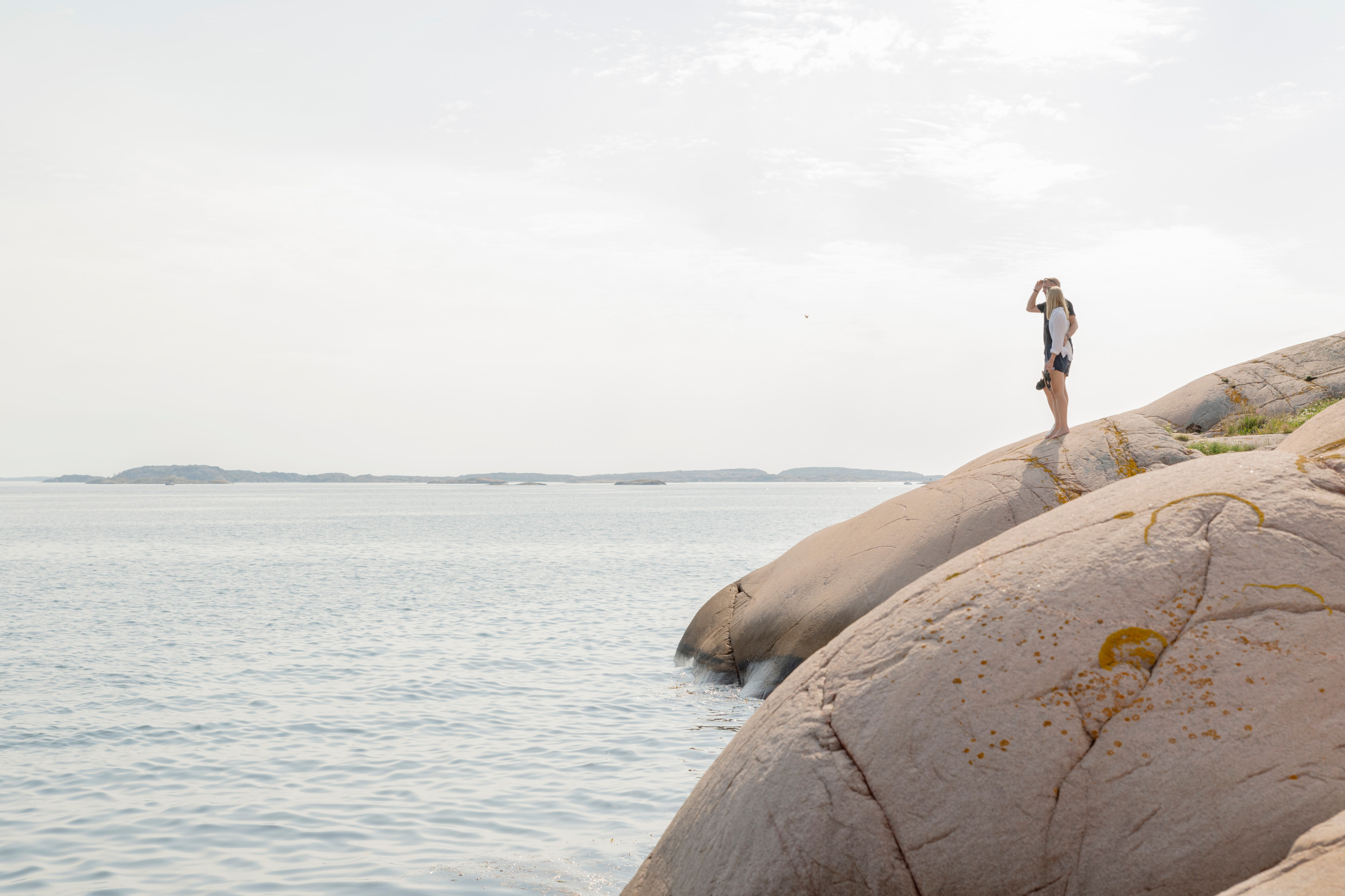 En man och en kvinna står på en klippa vid vattnet och tittar ut över skärgården. De är somrigt klädda och kvinnan har en kamera i handen.