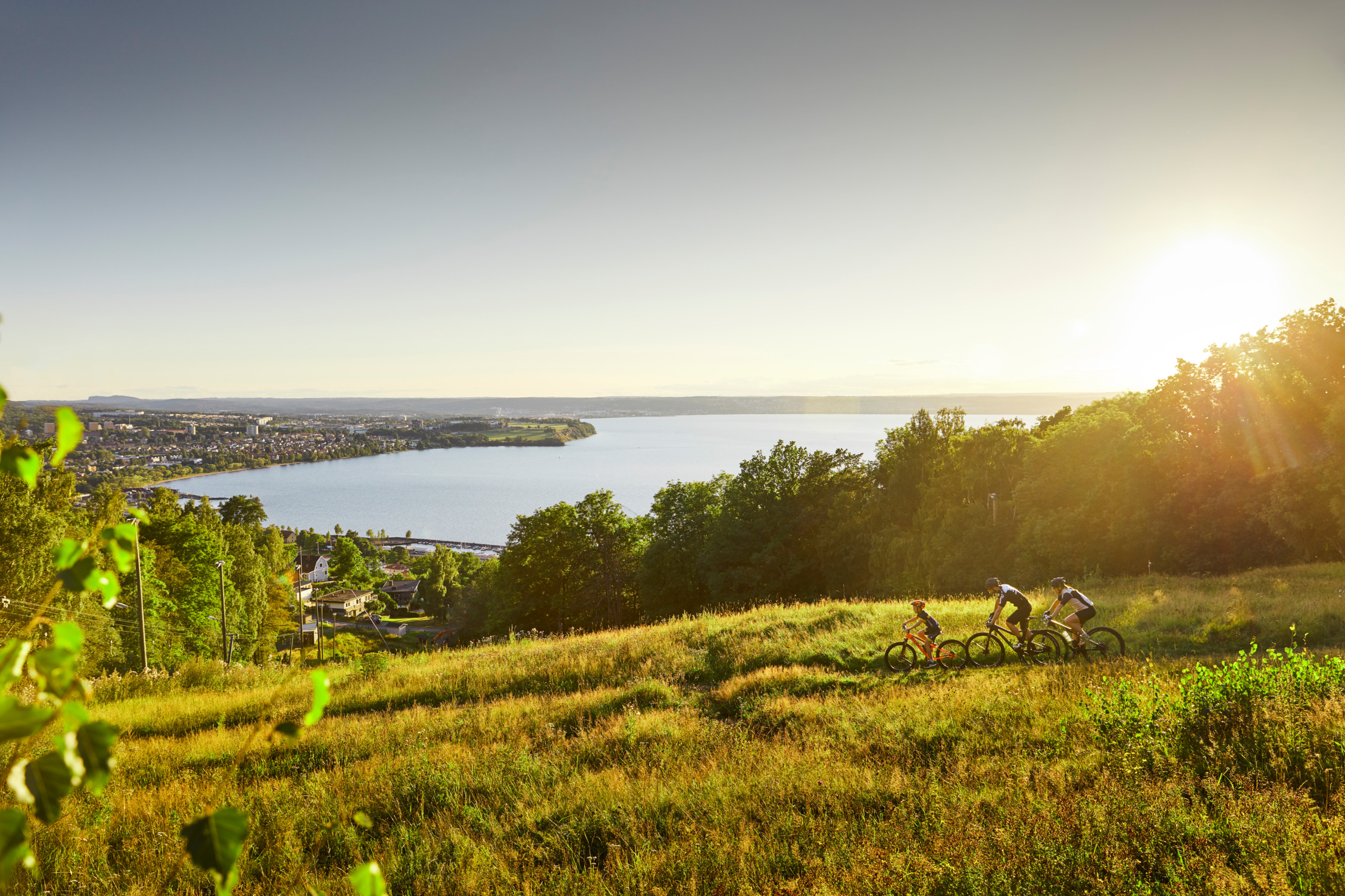Tre personer cyklar ner för en backe på gräs. Runtomkring finns en äng, skog och en i bakgrunden ser man en stad framför en sjö.