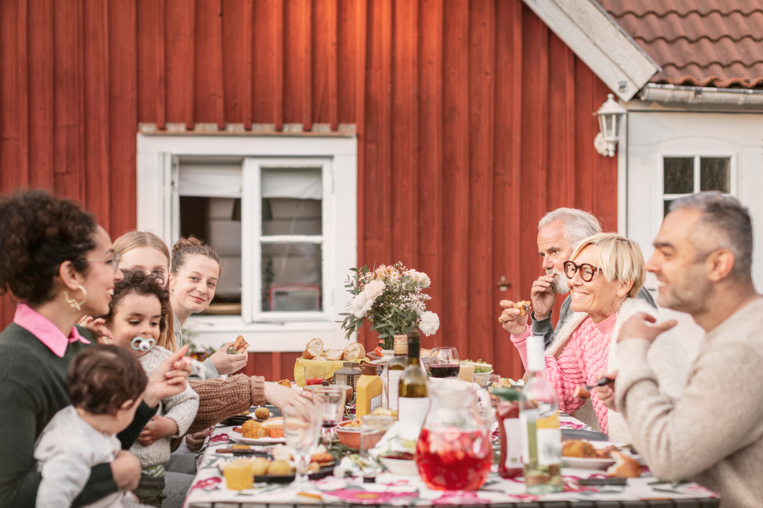 Ett långbord är uppdukat utomhus med en stort sällskap som sitter och äter grillad mat, bröd och dricker vin eller saft till maten.