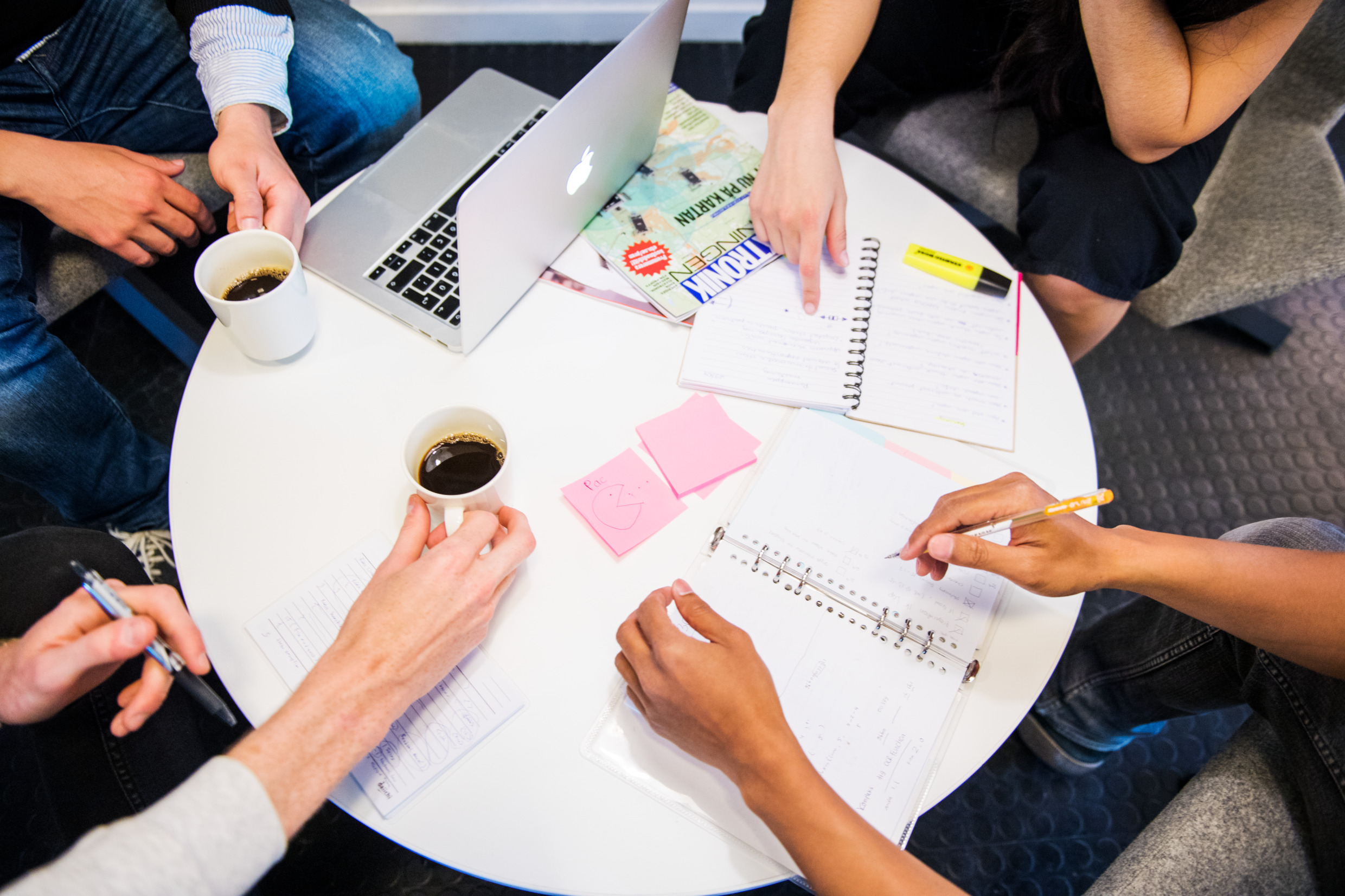 Ett bord med en dator, papper och kaffekoppar på. Bilden är tagen uppifrån och i bildens ytterkant sticker det in händer från alla sidor.