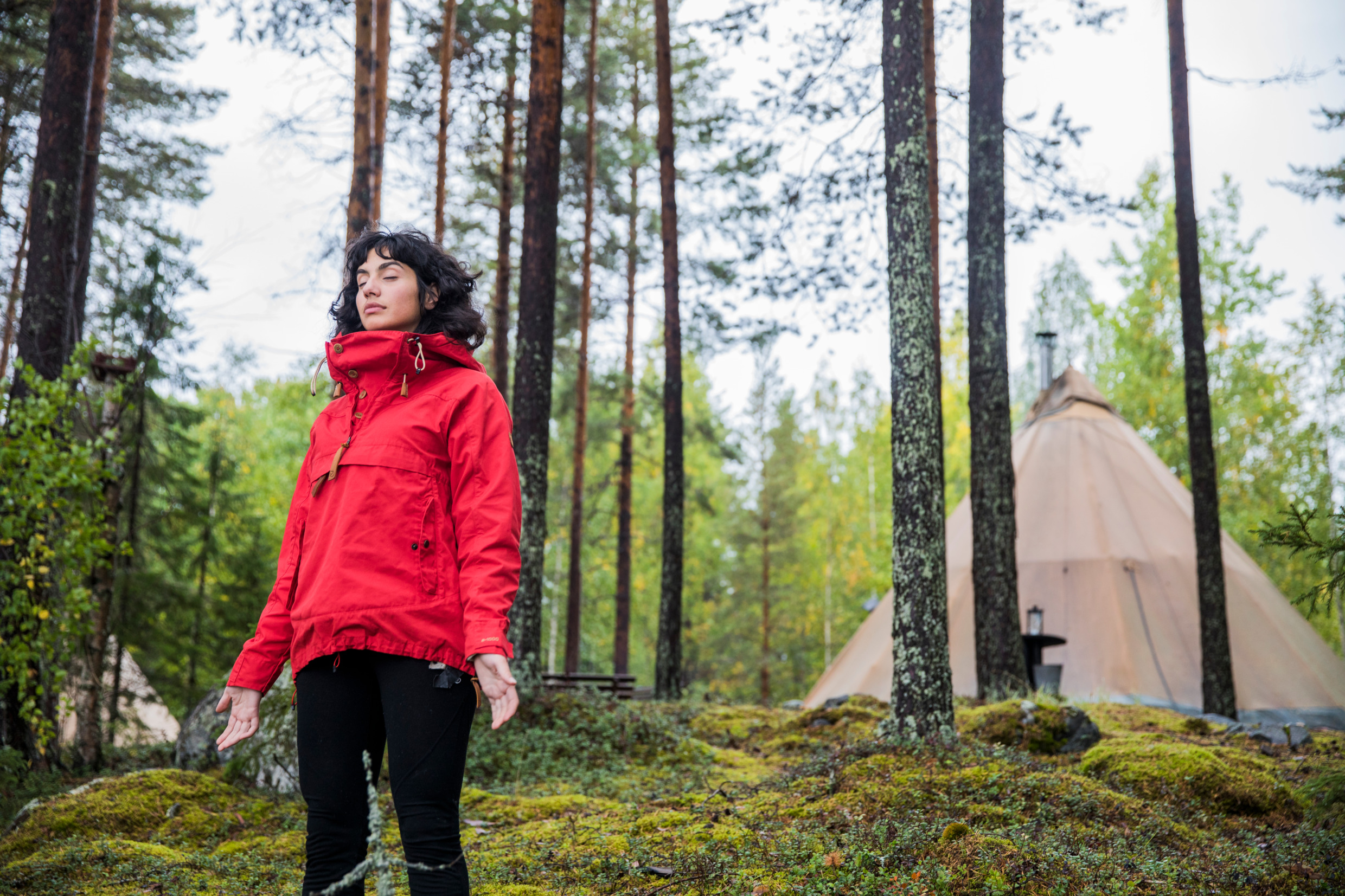 Kvinna i klarröd jacka i skogen står och blundar med händerna längs med sidorna och handflatorna riktade framåt. Bakom henne finns träd och ett tält är uppställt några meter bort.