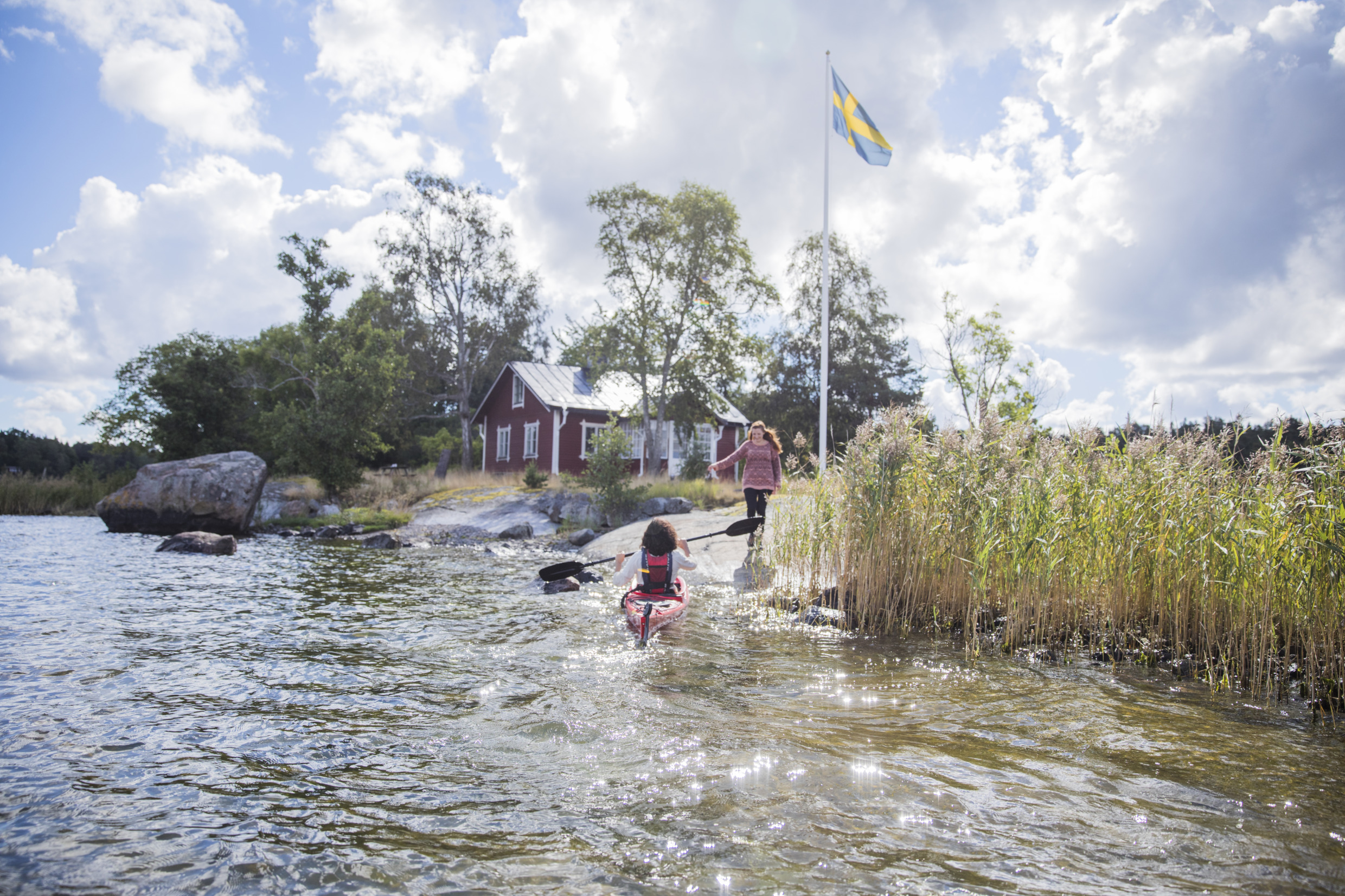 En flicka paddlar kajak upp mot klippkanten där en annan flicka kommer gående. Bakom dem står en svensk flagga hissad och ett rött hus med vita knutar framför skog.