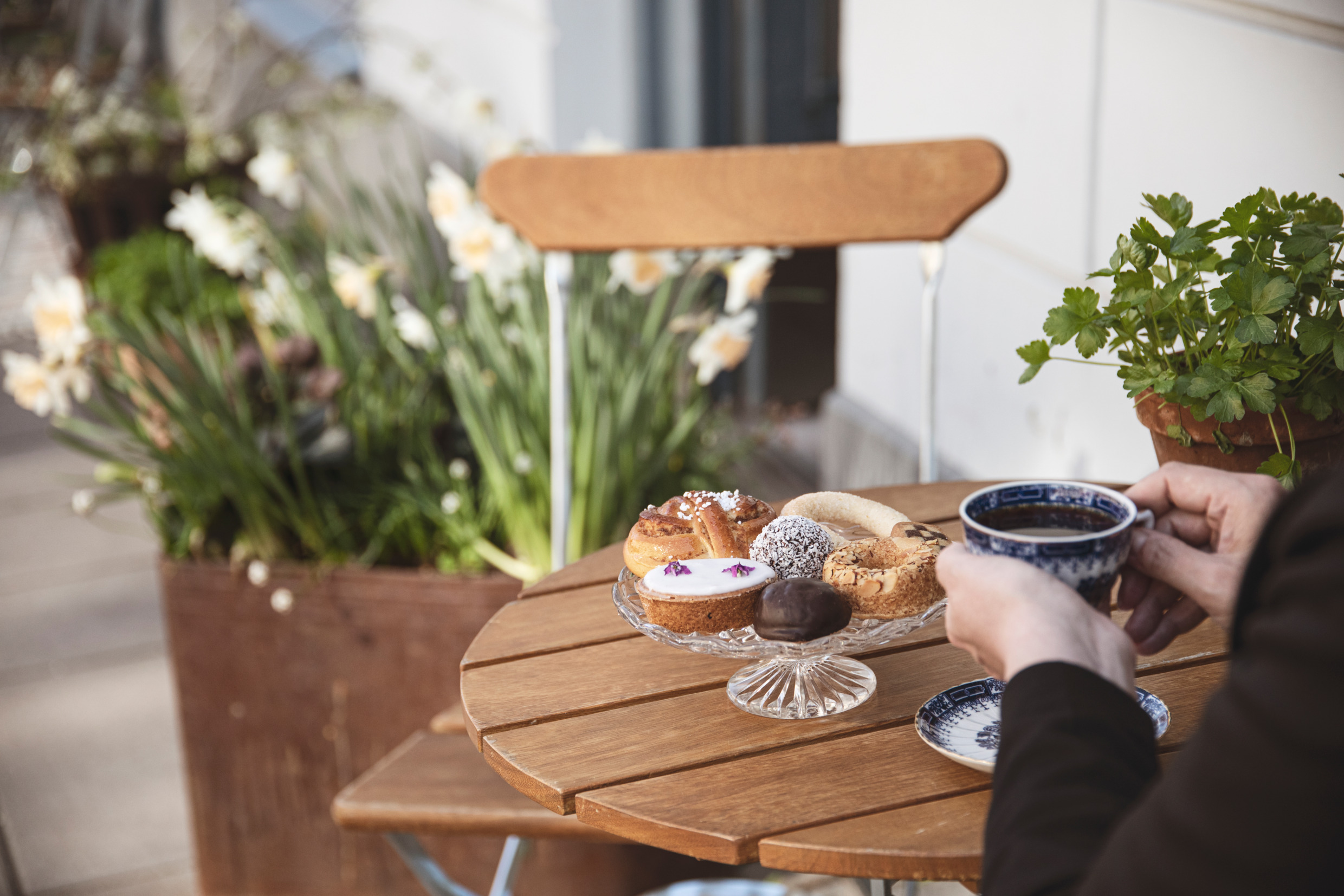En person sitter utomhus vid ett cafébord och dricker kaffe och har flera kakor och bullar på ett fat framför sig.