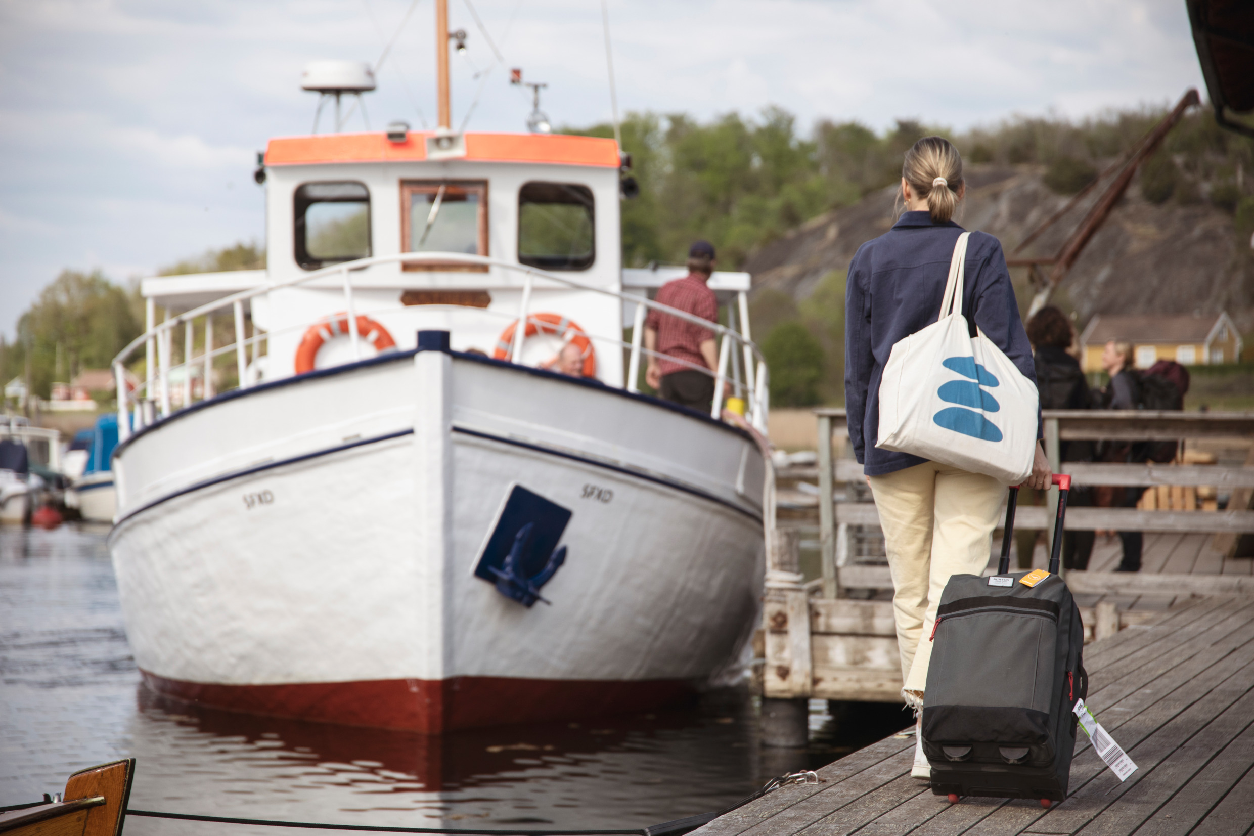 En kvinna går på en kaj och drar en väska efter sig. På kajen står en skärgårdsbåt och väntar på henne.