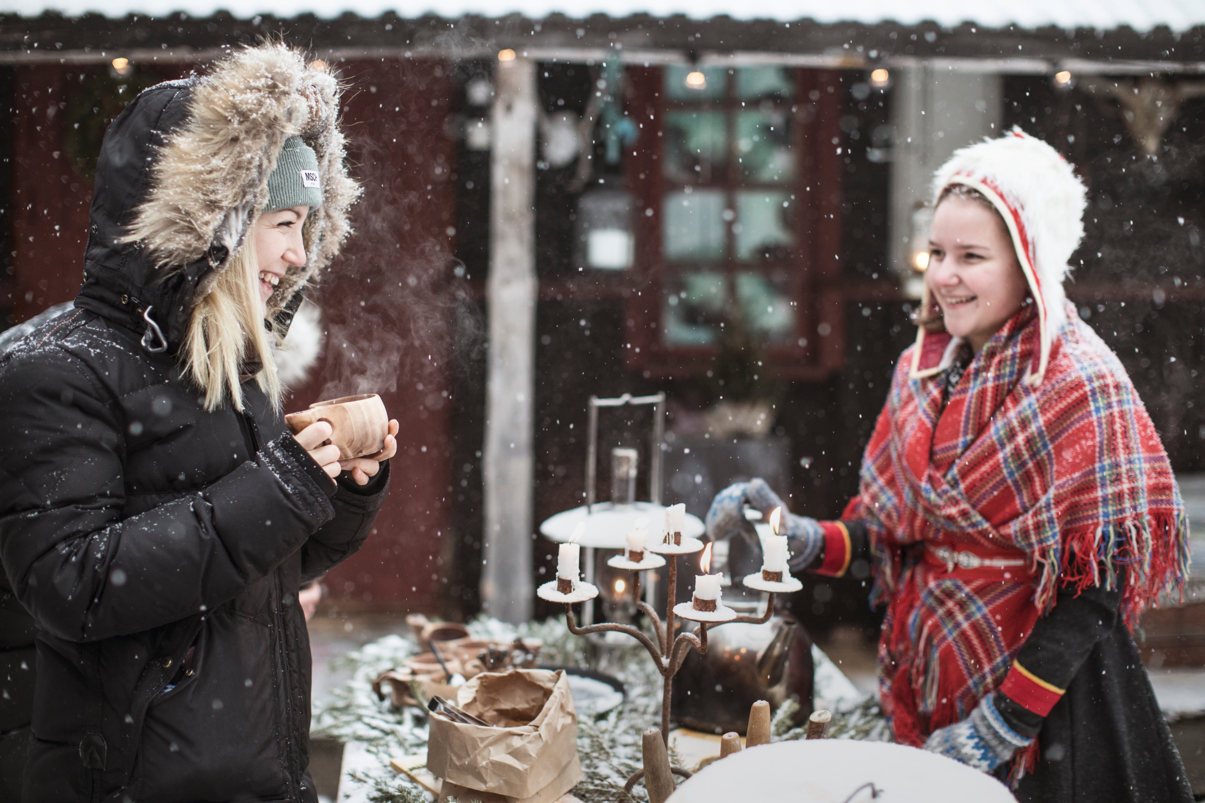 Två personer står och fikar runt ett bord utomhus medan snön singlar ner från himlen.