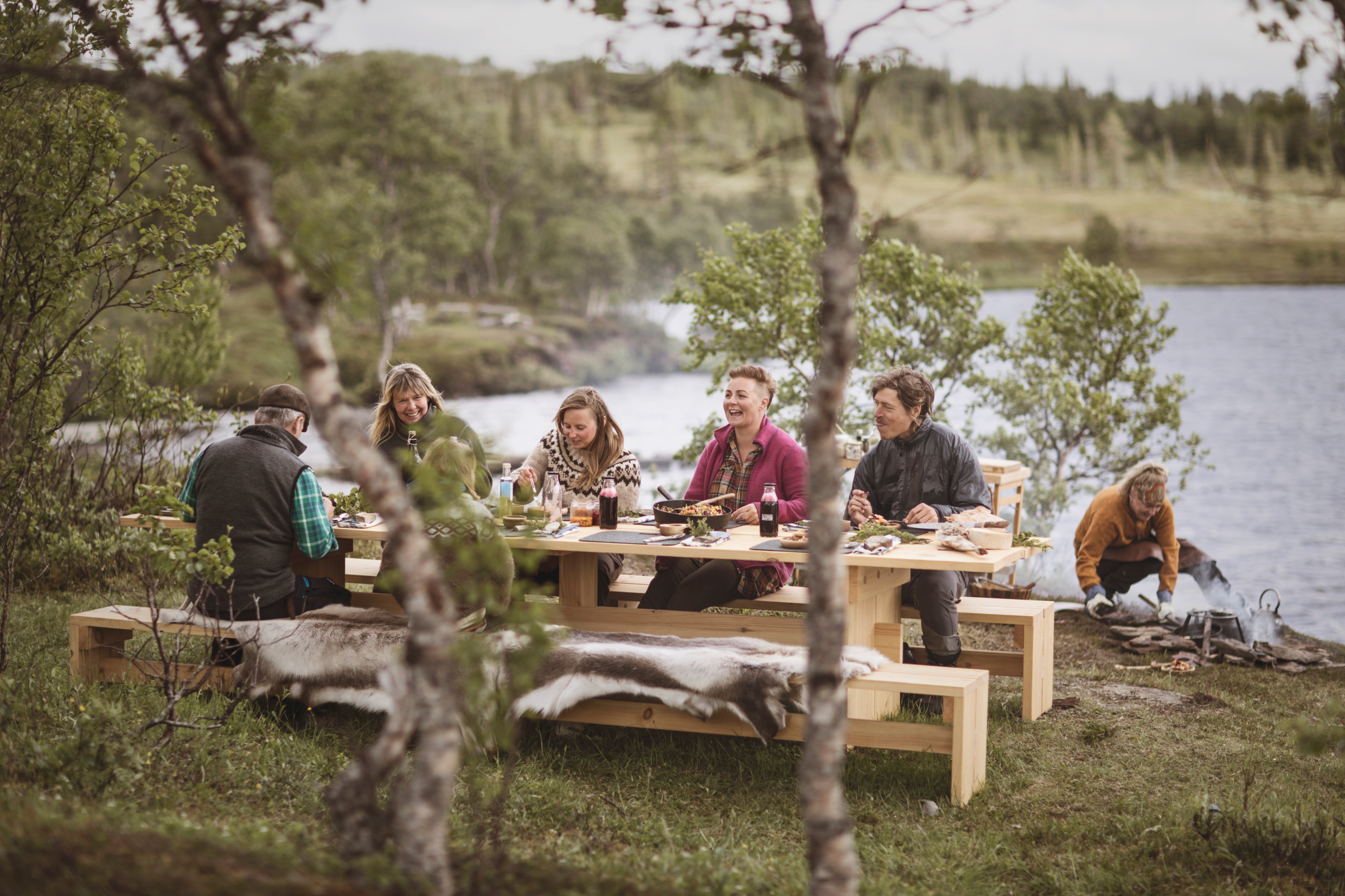 Fem personer sitter runt ett stort, grovt bord av träd mitt i björkskogen. I bakgrunden håller en person på att laga mat över öppen eld.