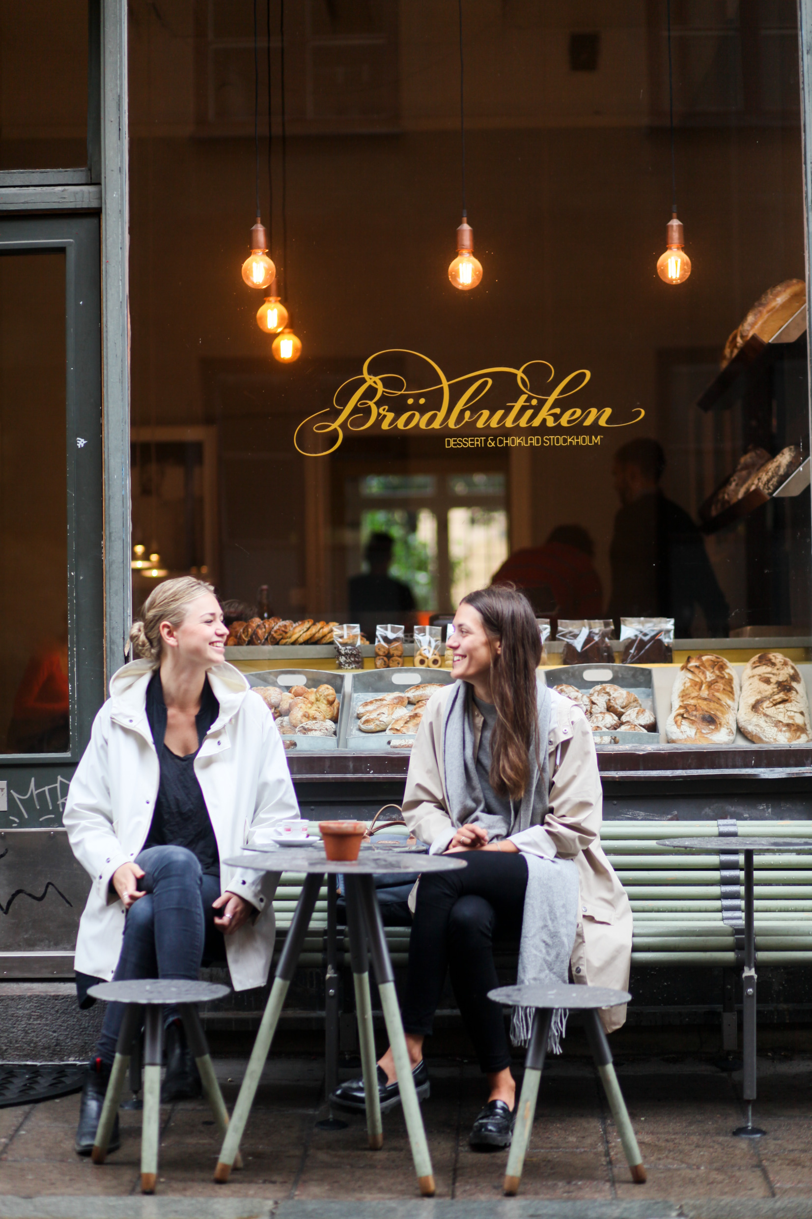 Två kvinnor sitter vid en liten uteservering utanför en brödbutik.