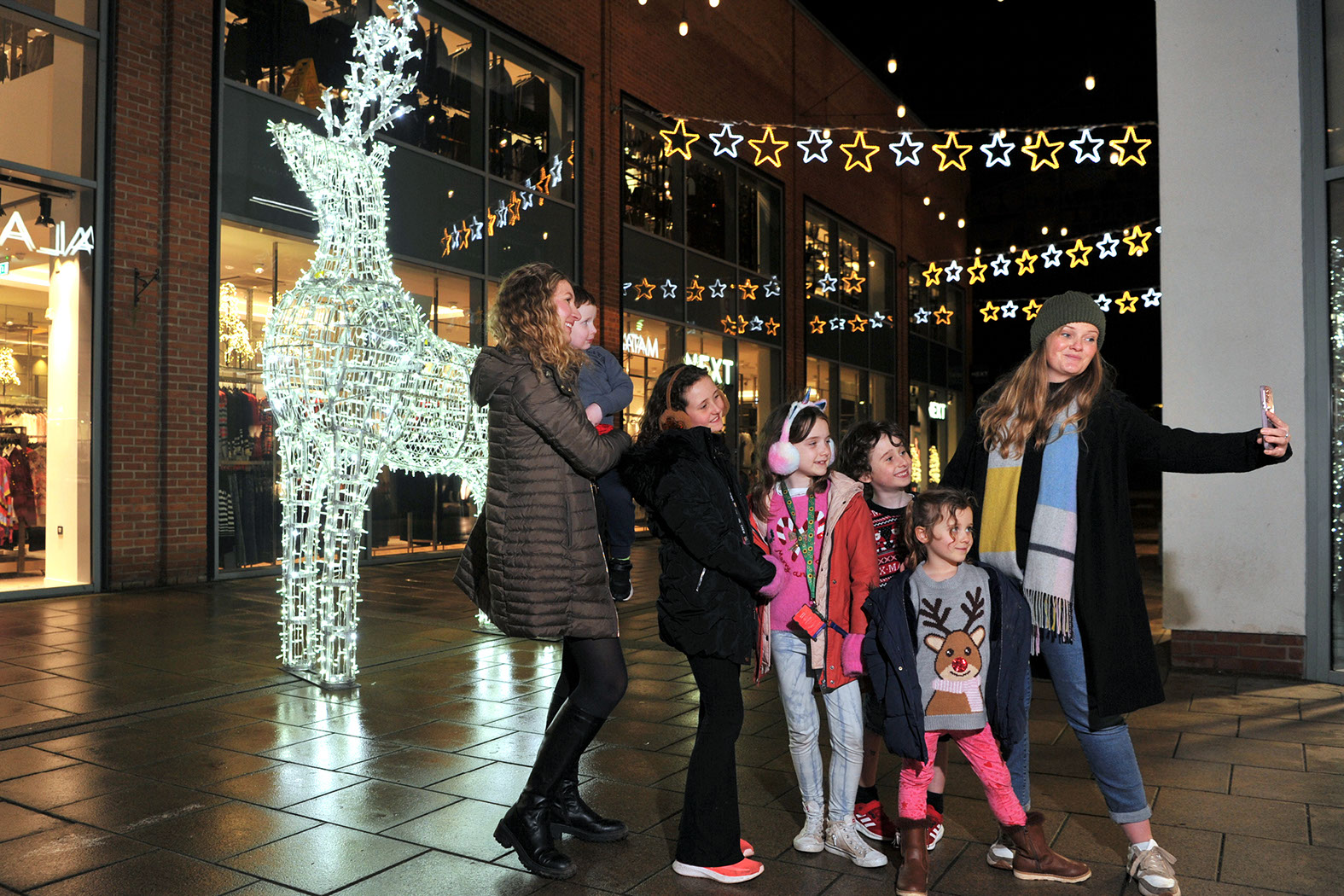 Two women with young families taking a group selfie in front of a large illuminated 3D reindeer